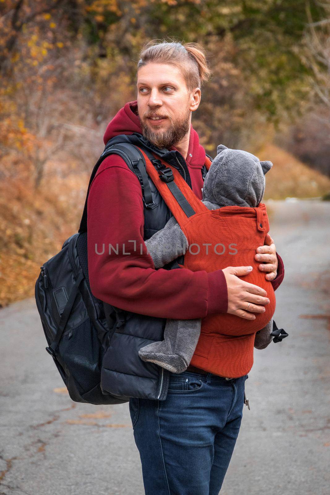 Young bearded hipster babywearing father with his baby in sling fall walking outdoor.