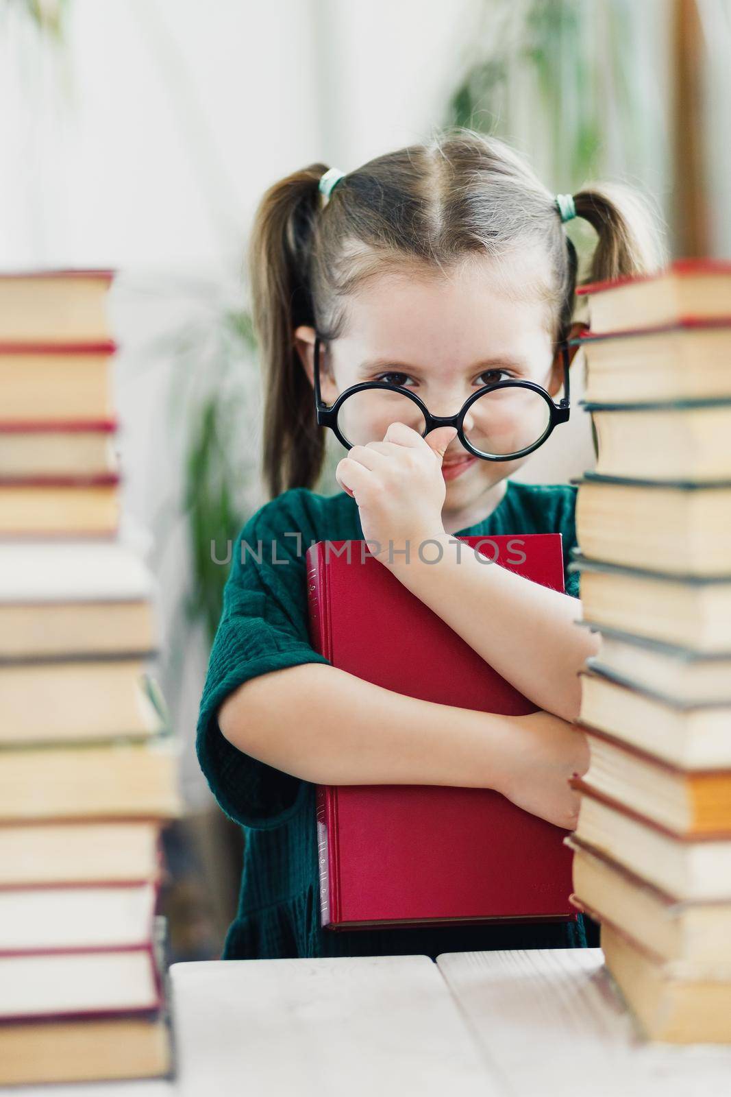 Cute little girl in green dress with red book in her hands fixing a glasses on her nose. Reading and education concept.
