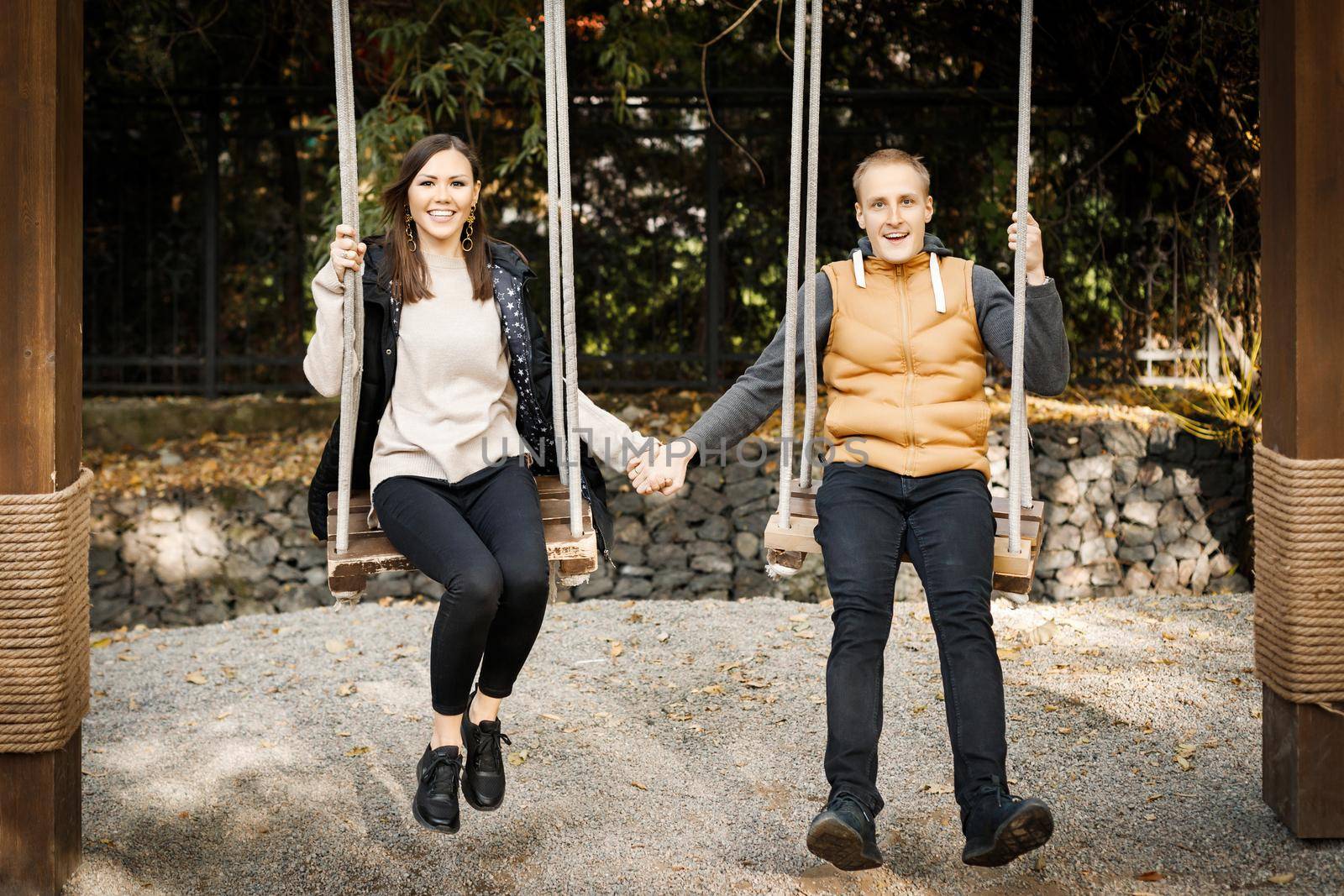Young couple laughing holding hands on a swing in the forest among the trees. High quality photo