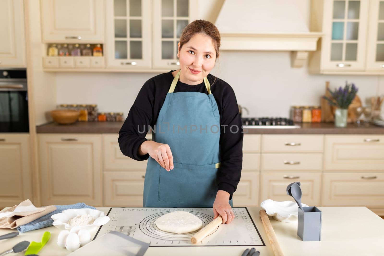 Asian woman cook in apron look at camera sprinkles flour on cooked wheat pastry dough on silicone baking mat in modern kitchen.