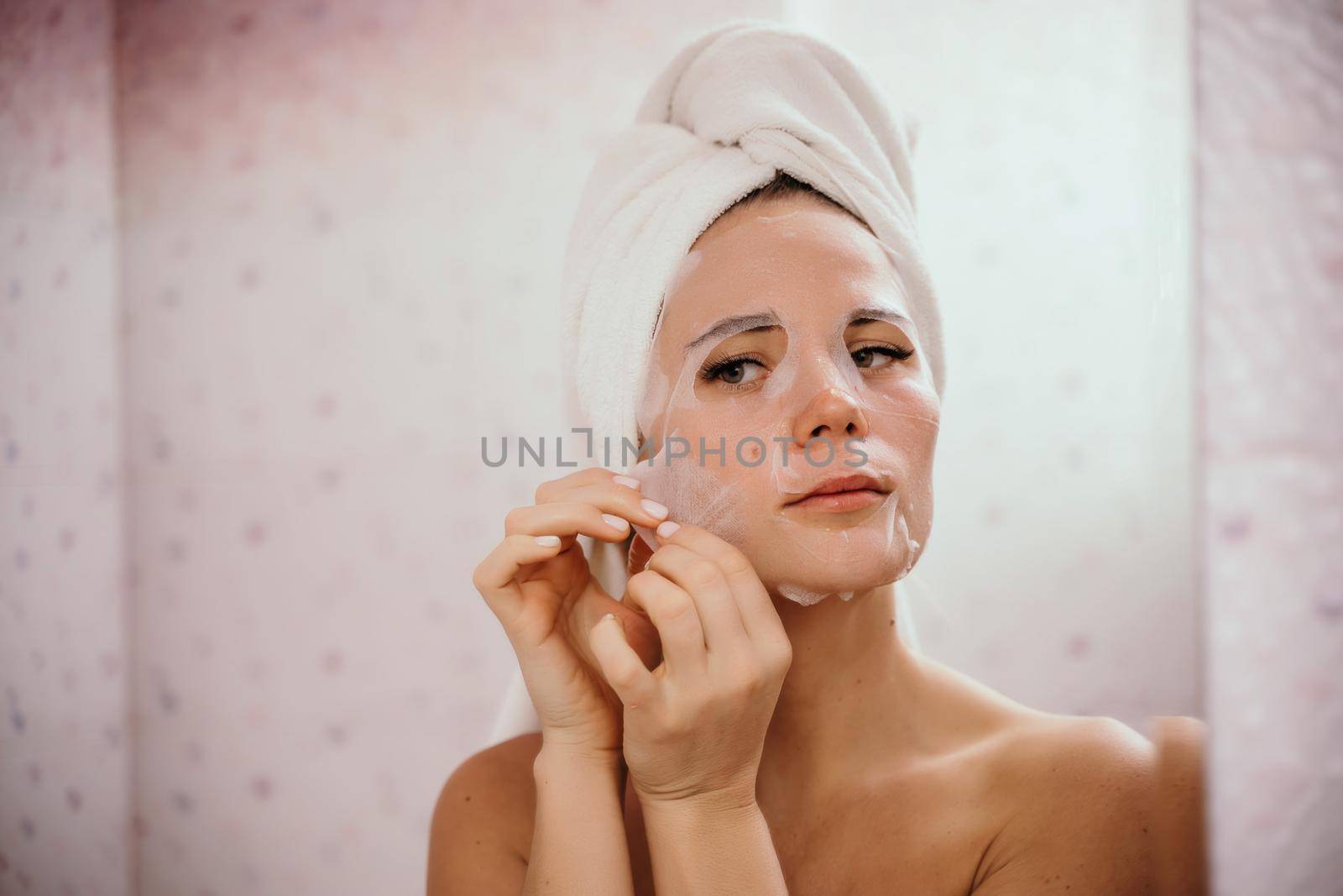Young beautiful woman using a moisturizing facial mask after taking a bath. Pretty attractive girl in a towel on her head stands in front of a mirror in a home bathroom. Daily hygiene and skin care by Matiunina
