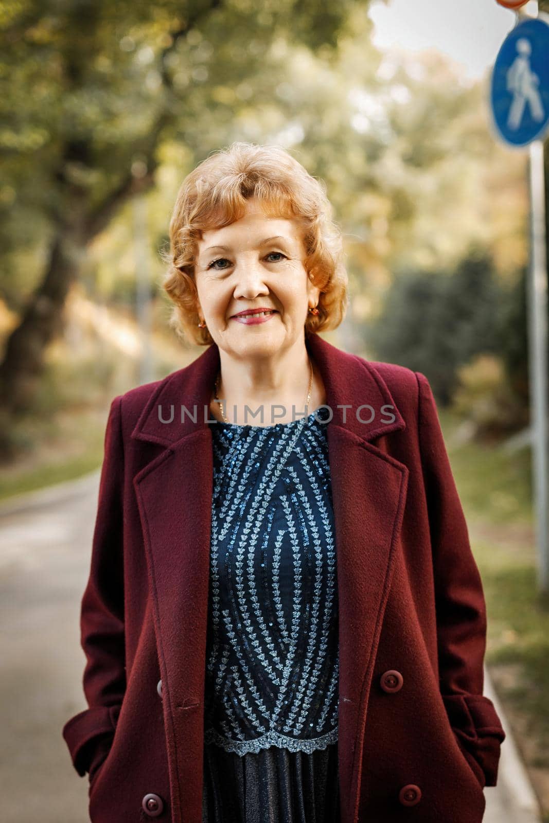 Mature happy pensioner woman stands outdoors in autumn in an unbuttoned dark color coat, smiles looking at the camera.