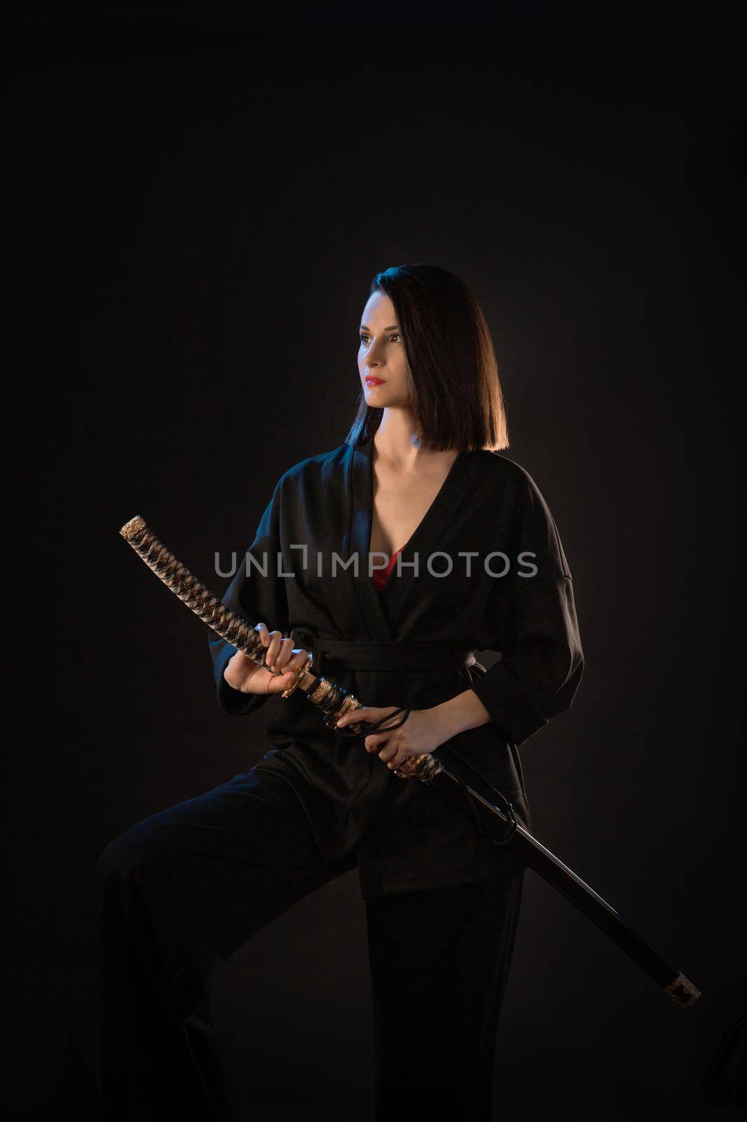 Young beautiful woman in black kimono with katana on black background.