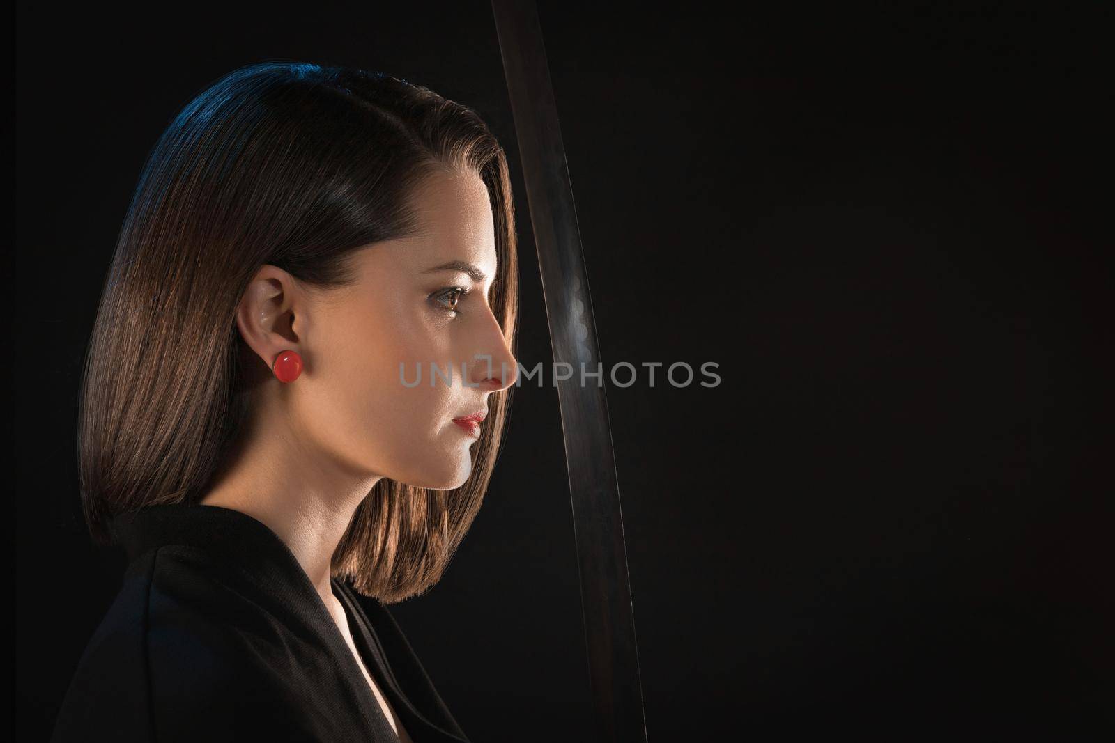 Portrait in profile of a young woman with red lips and earrings in front of a sharp blade of a saber on a black background.