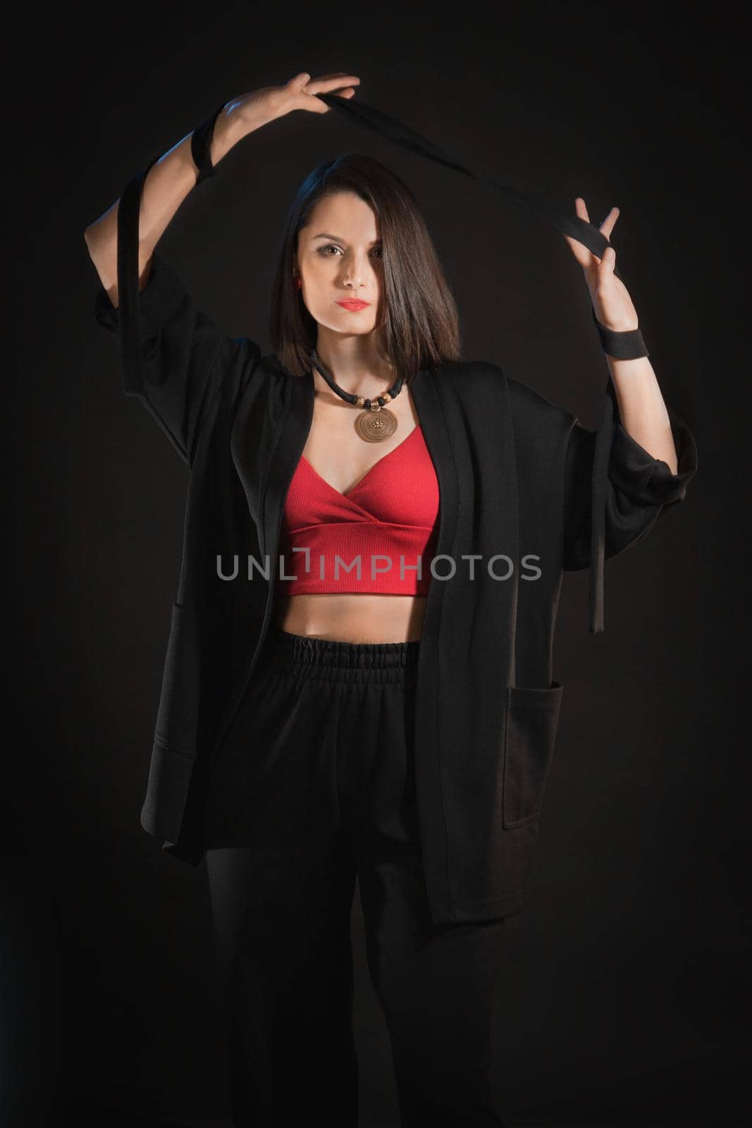 Fashion studio shot in low key with woman with belt in raised hands in black sleepwear.