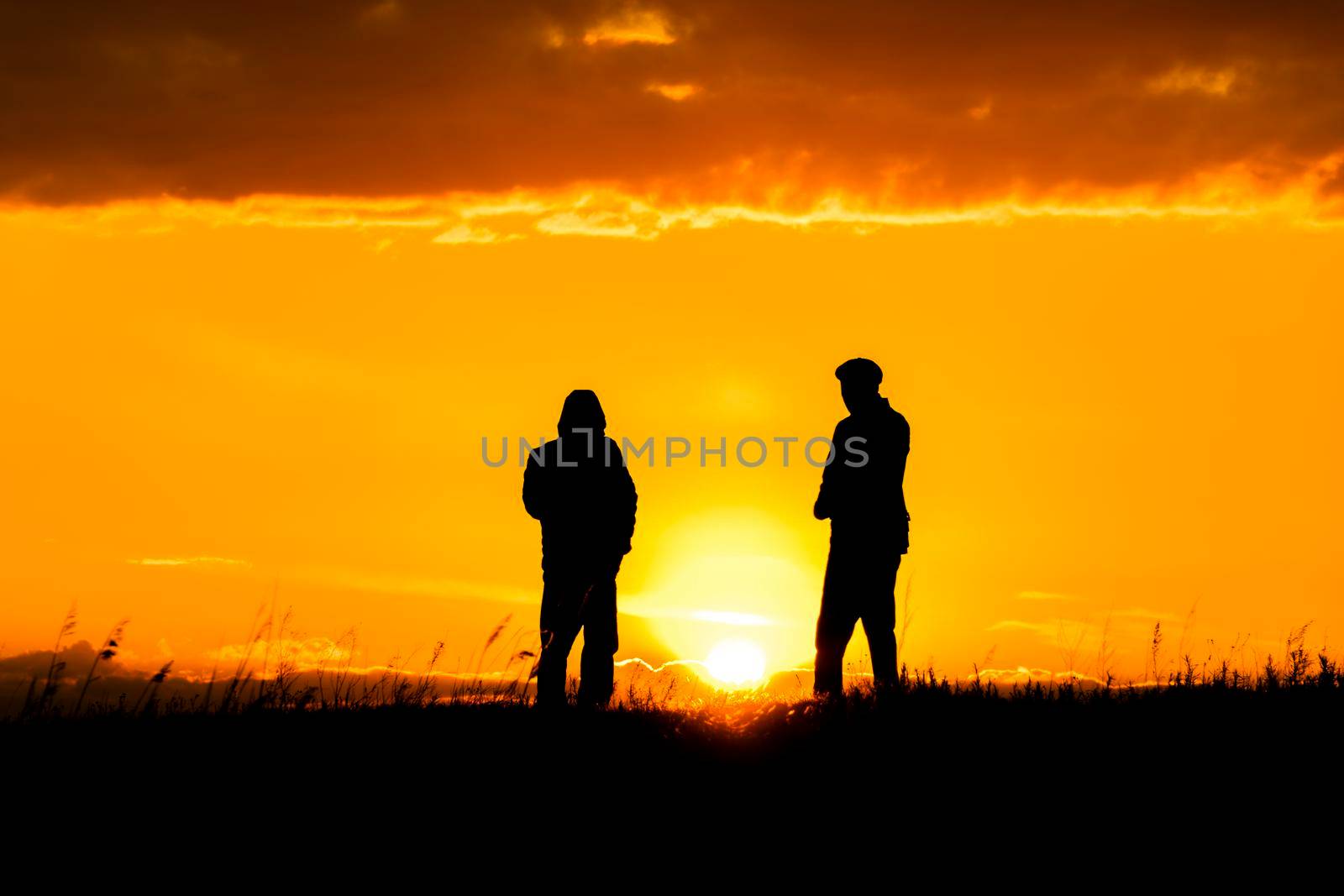 Two silhouettes of men talking at sunset or sunrise with dramatic sky and clouds.Dialogue and meeting two people on the horizon and skyline.Outlines of people in the sunlight.Golden hour with friends by YevgeniySam