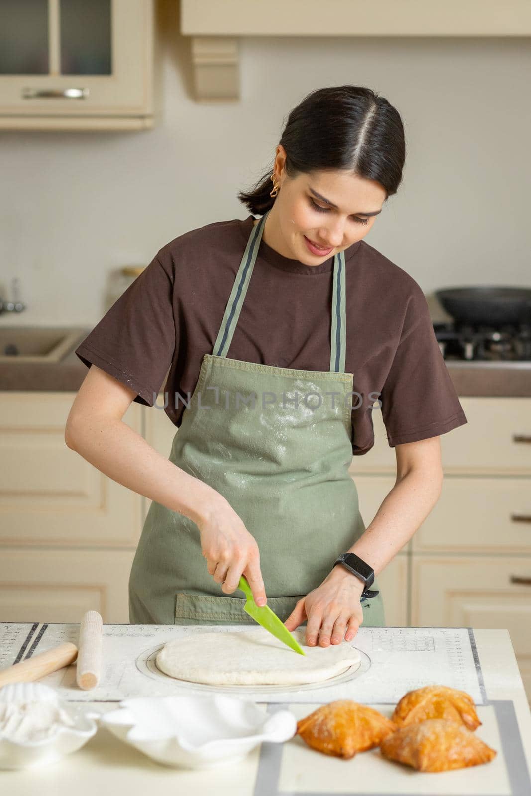 Young brunette woman cook in green apron cuts rolled out dough with plastic knife on silicone baking mat.