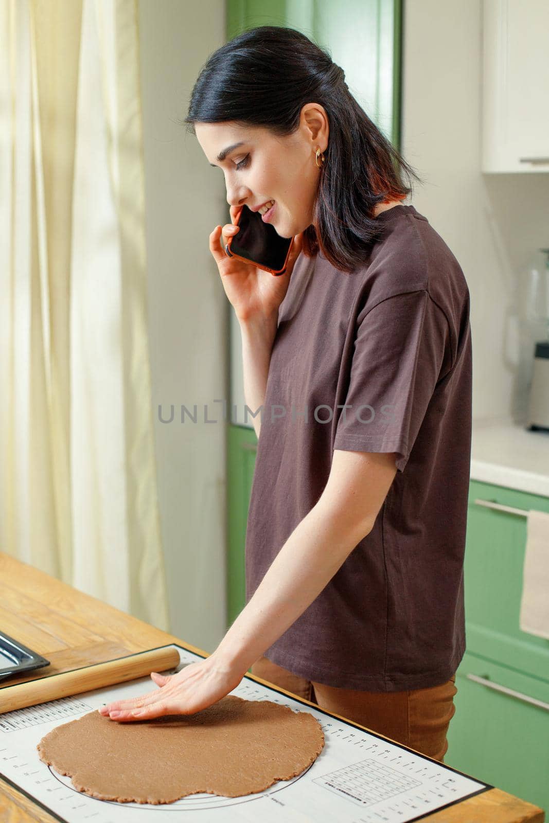 Vertical portrait of young mixed race happy woman talking on smartphone in home kitchen while cooking.