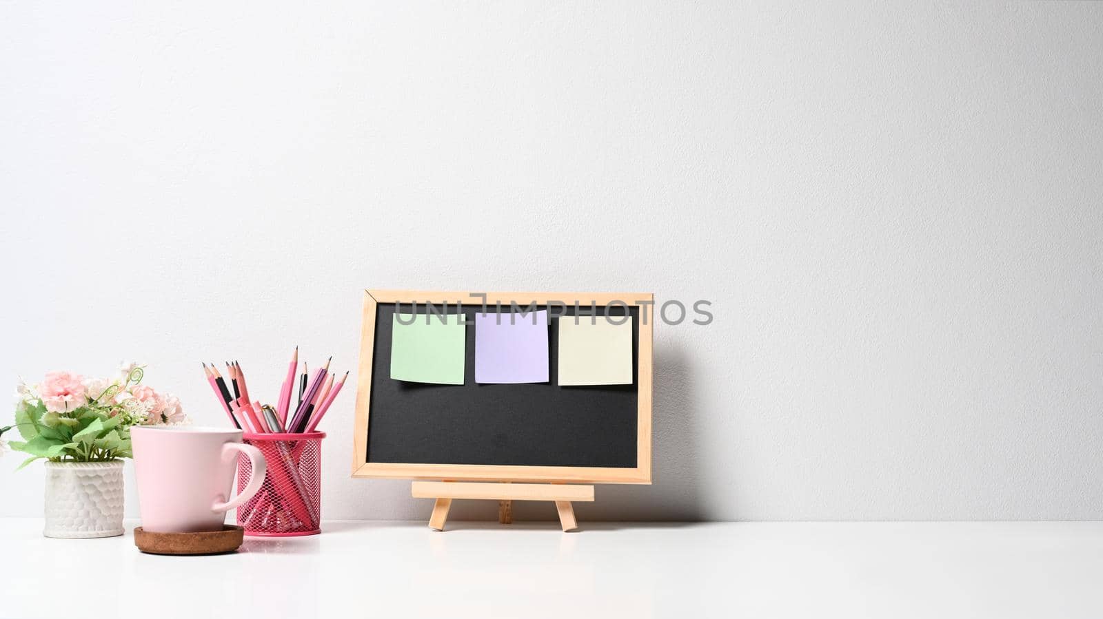 Black board with colorful sticky notes, pencil holder, coffee cup and flower pot on white table by prathanchorruangsak