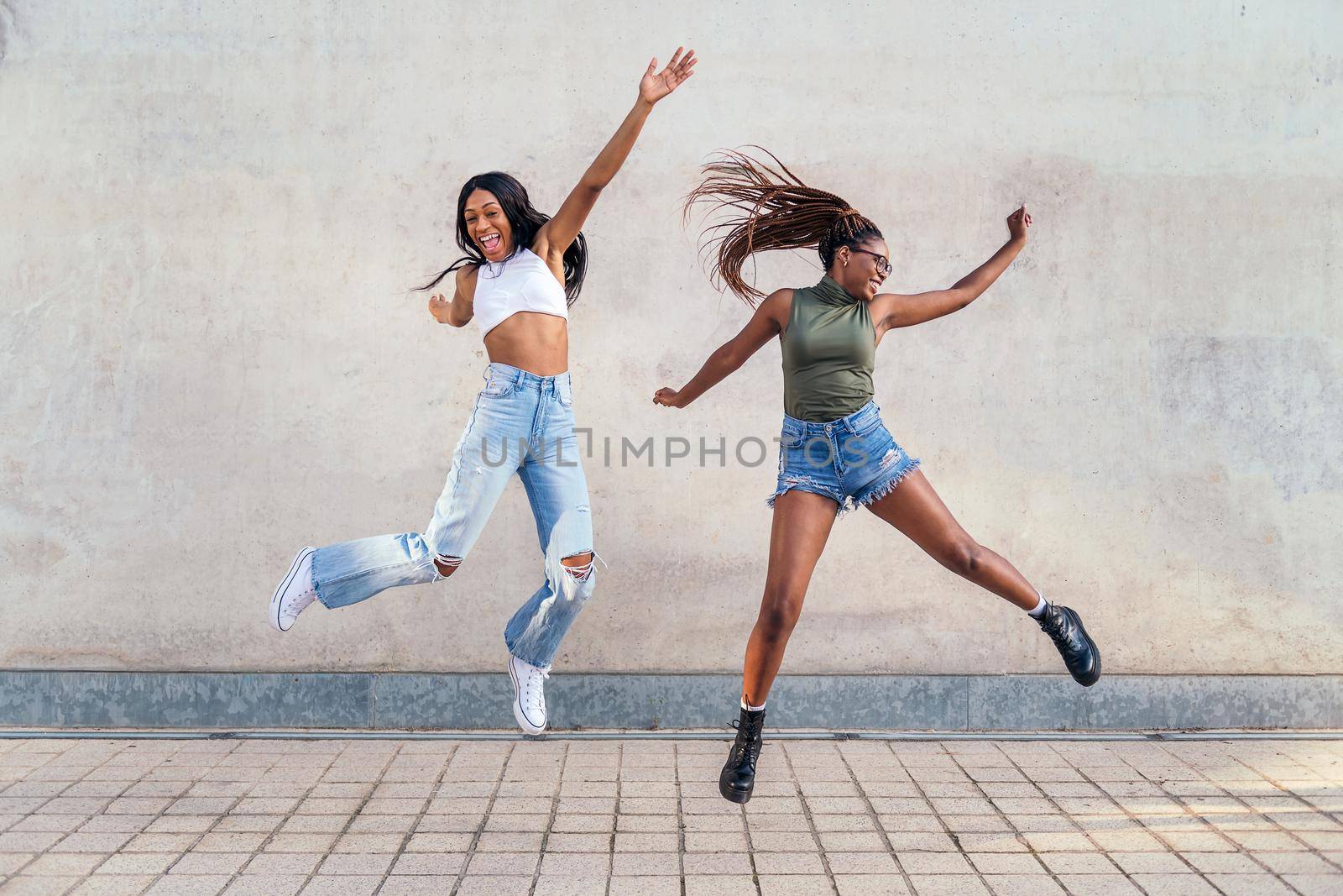 two young black girls jumping happy with a gray concrete wall in the background, concept of friendship and urban lifestyle