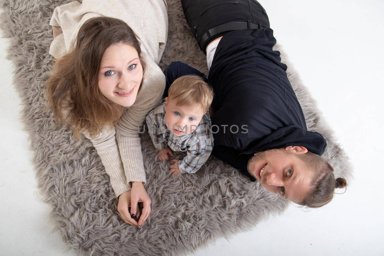 A young caucasian family with a child lies on a carpet on a white floor and looks at the camera. Top view.