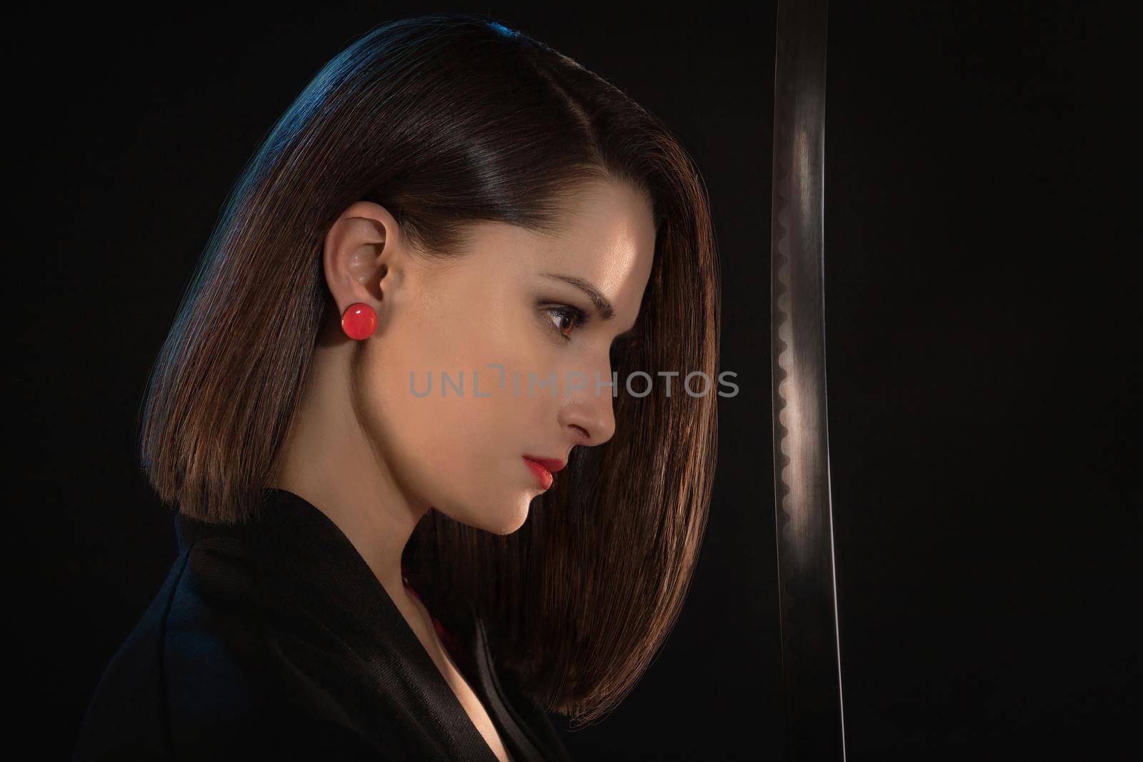 Portrait of a young woman with red lips and earrings in front of a sharp blade on a black background.