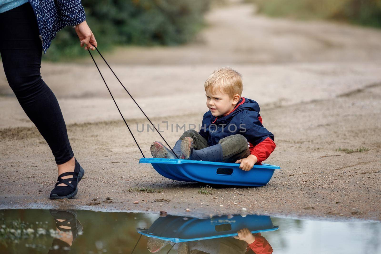 Cute boy playing with sled in summertime. Mother towing son on sleigh, portrait.