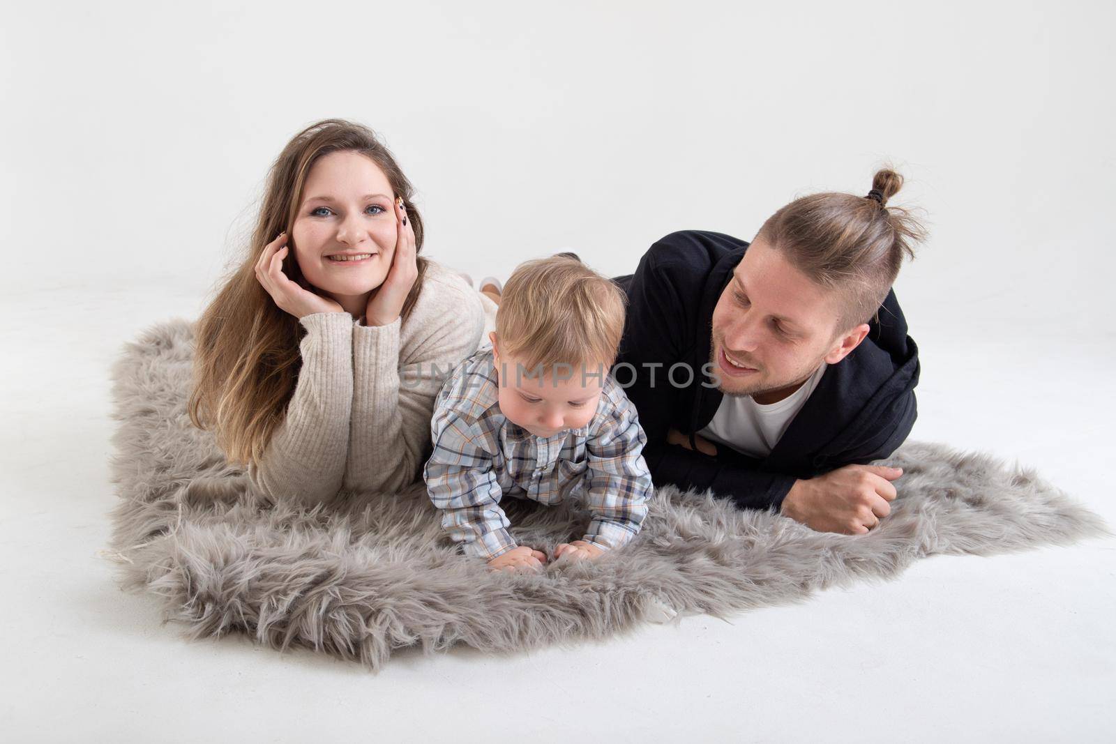 Young caucasian parents with one year old son lie on a carpet in a white studio.