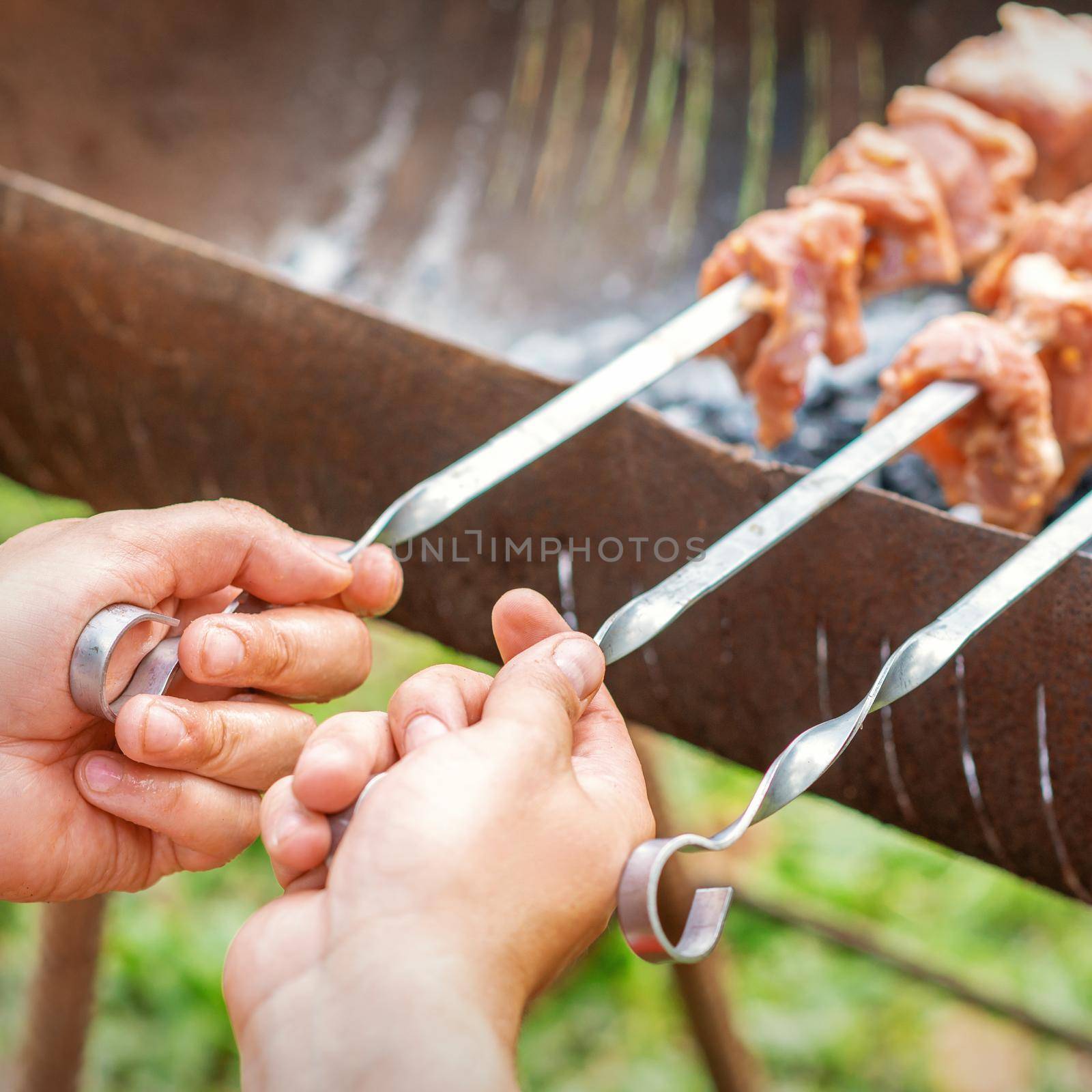 Hands of man prepares barbecue meat on skewer by grill on fire outdoors