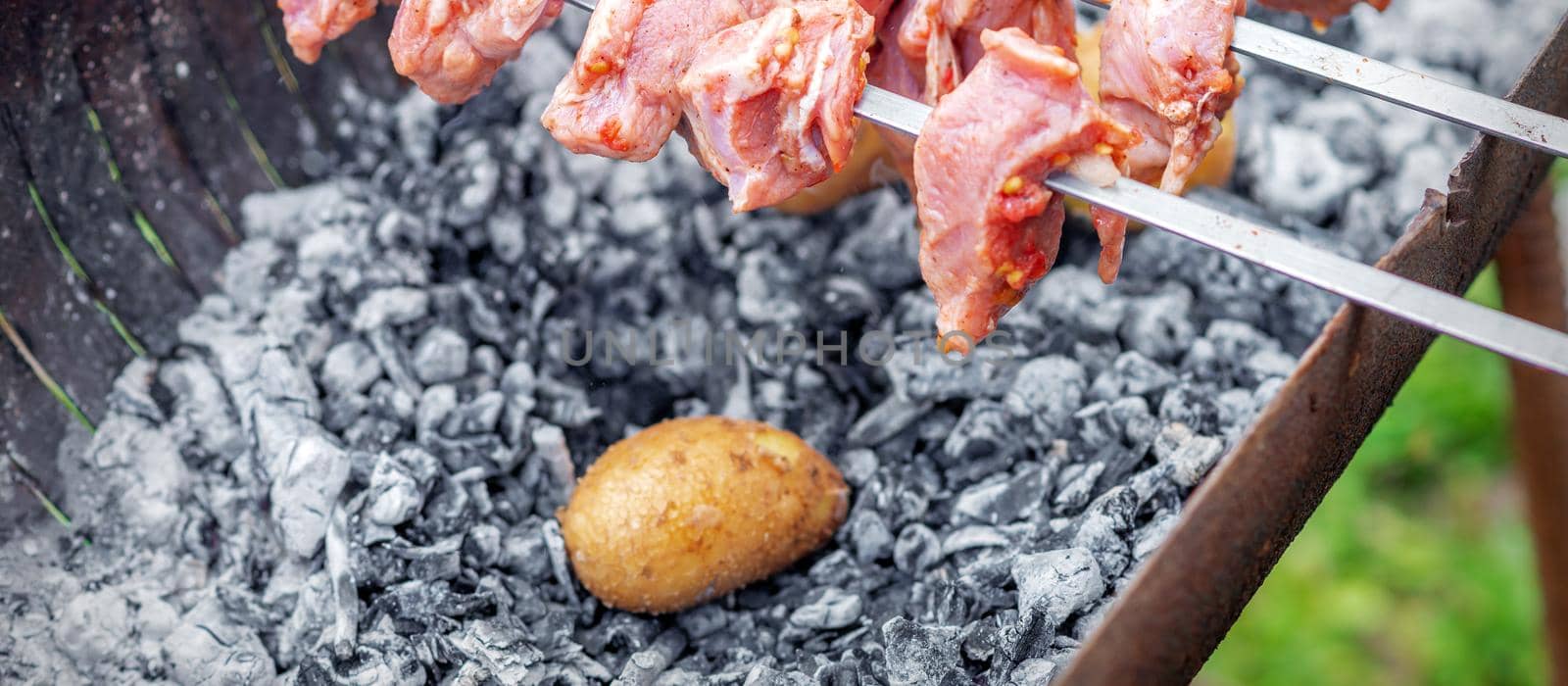 Hands of man prepares barbecue meat with potatoes on skewer by grill on fire outdoors. Concept of lifestyle rustic food preparation