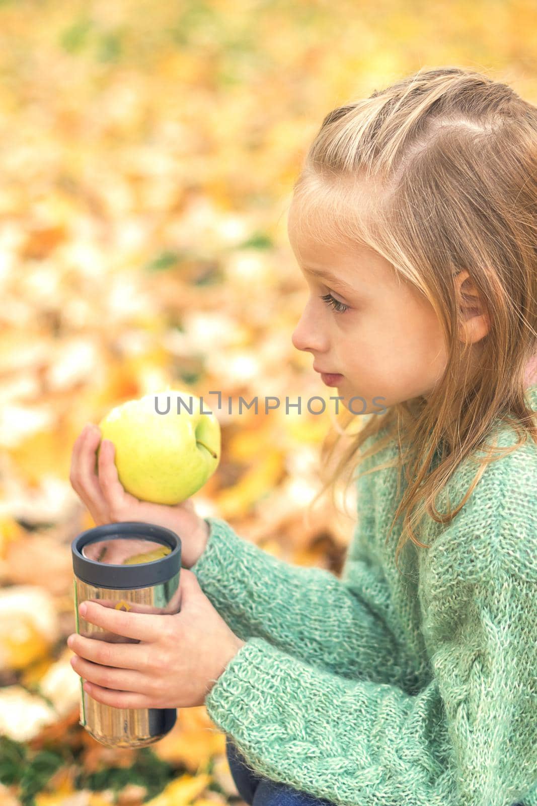 Beautiful little caucasian girl with apple and drink sitting in autumn park
