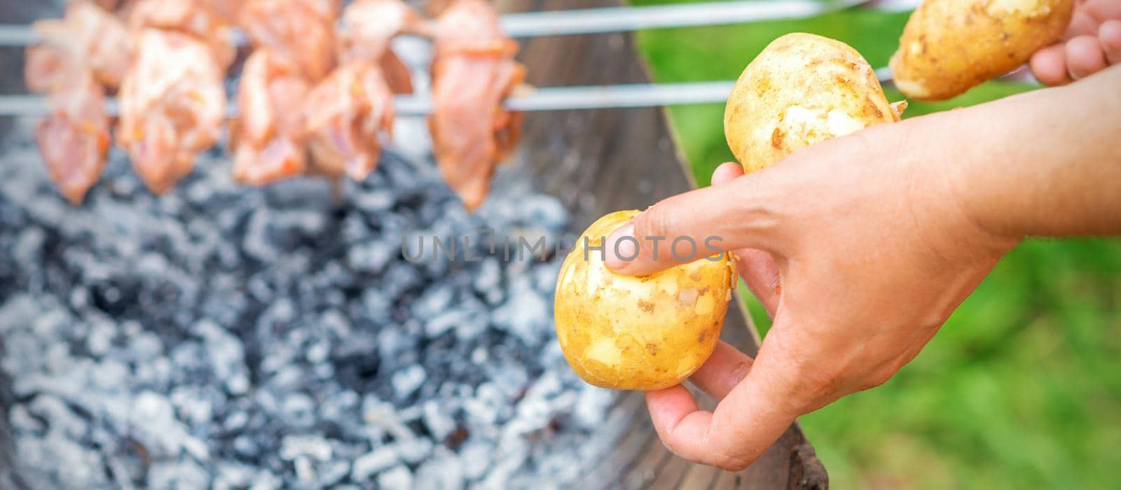 Hands of man prepares barbecue meat with potatoes on skewer by grill on fire outdoors. Concept of lifestyle rustic food preparation