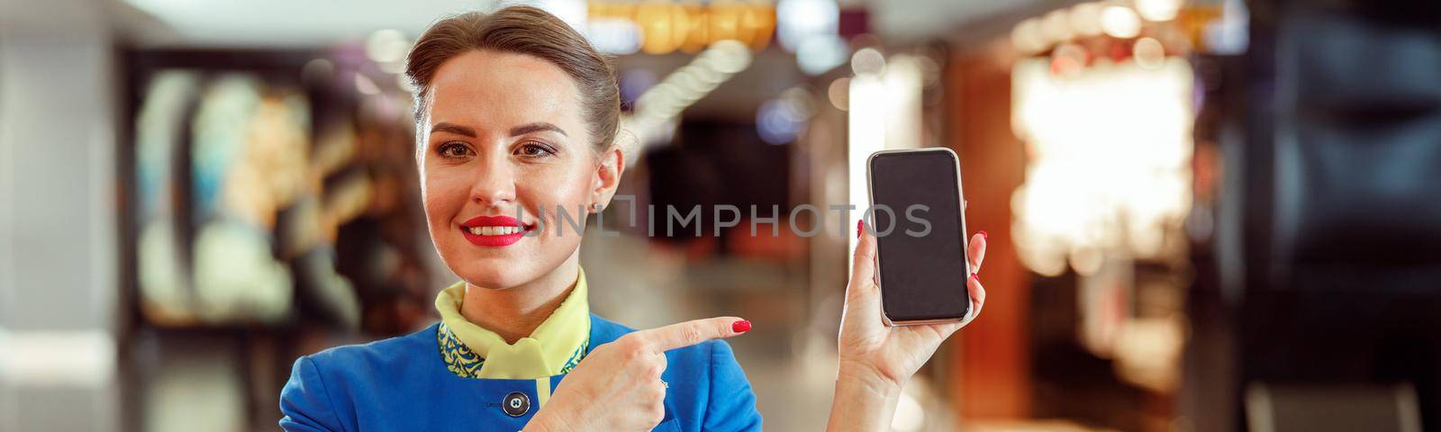 Cheerful stewardess in aviation air hostess uniform holding cellphone and smiling while standing in airport terminal