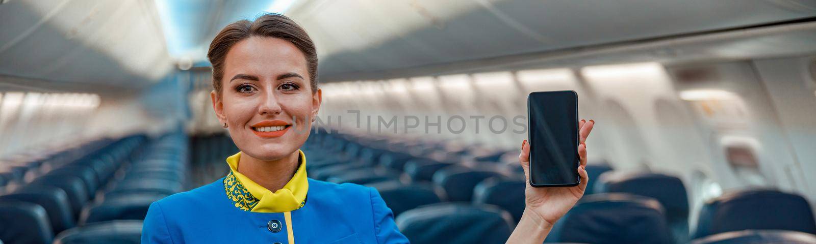 Smiling woman flight attendant holding mobile phone and pointing at passenger seat while standing in aircraft salon