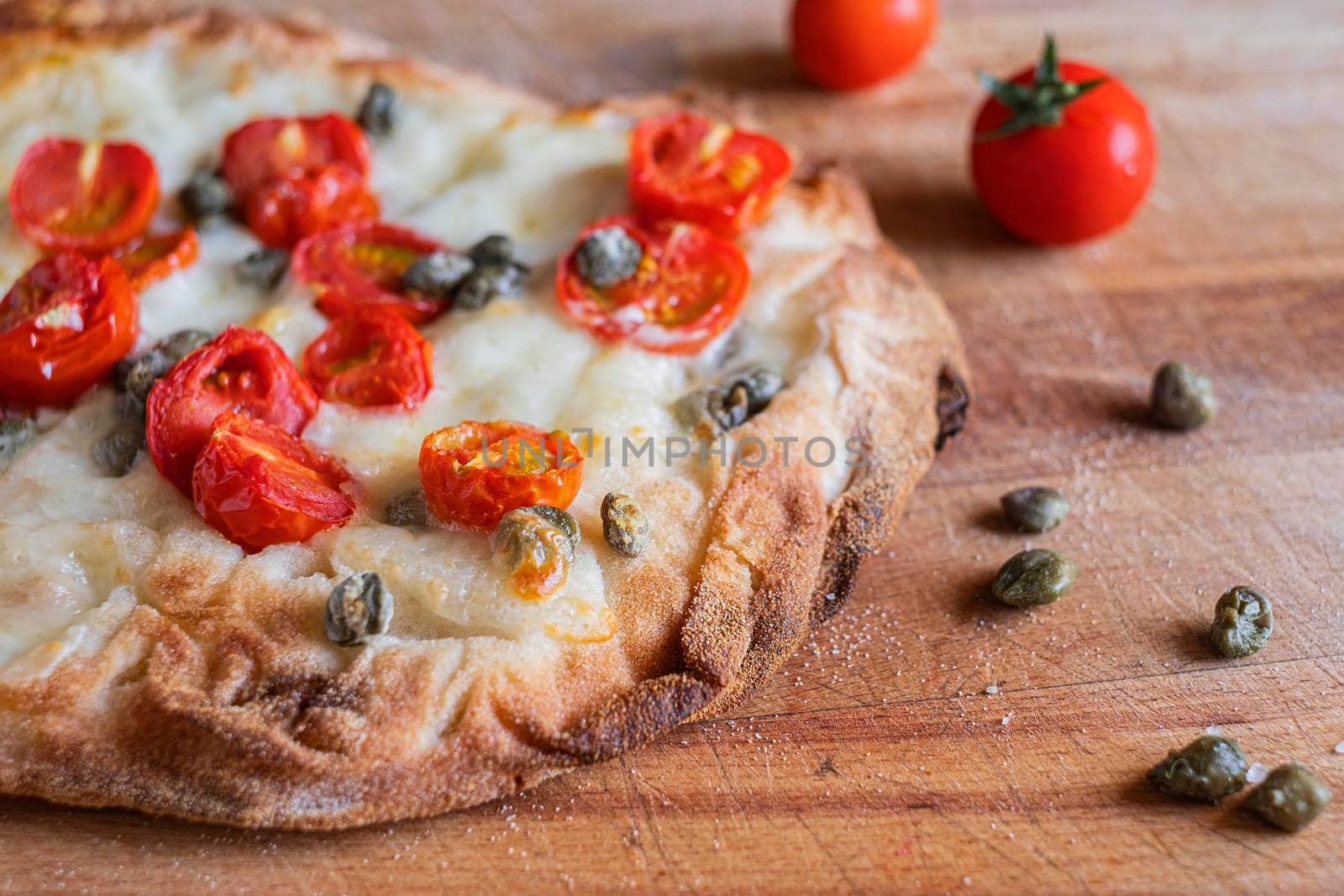 Small homemade vegetarian pizza with addition of tomatoes, capers on a wooden rustic table.