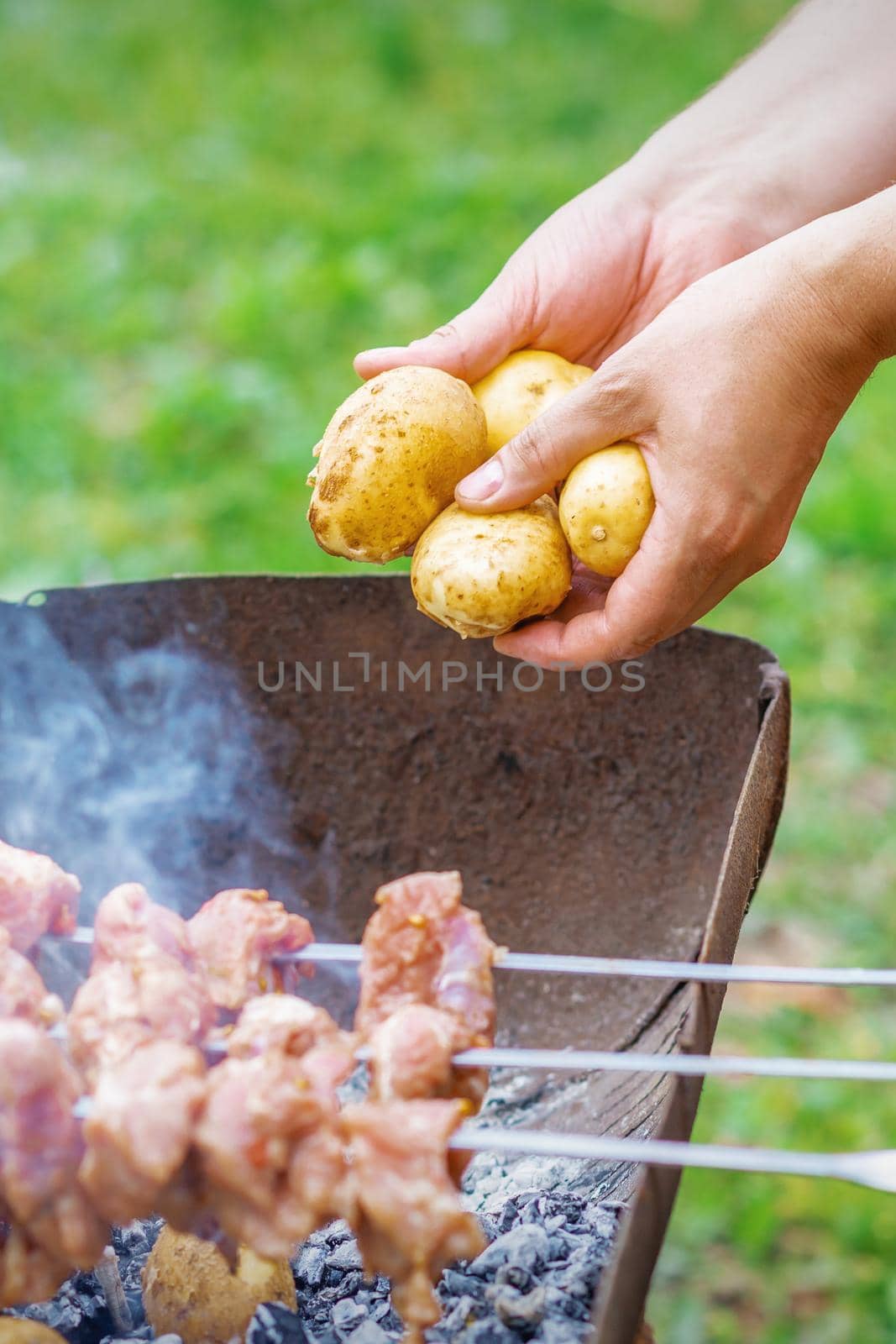 Hands of man prepares barbecue meat with potatoes on skewer by grill on fire outdoors. Concept of lifestyle rustic food preparation