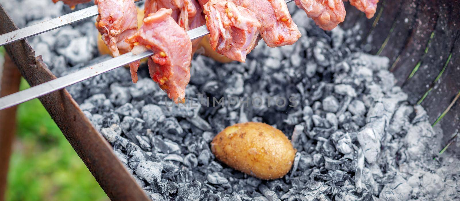 Hands of man prepares barbecue meat with potatoes on skewer by grill on fire outdoors. Concept of lifestyle rustic food preparation