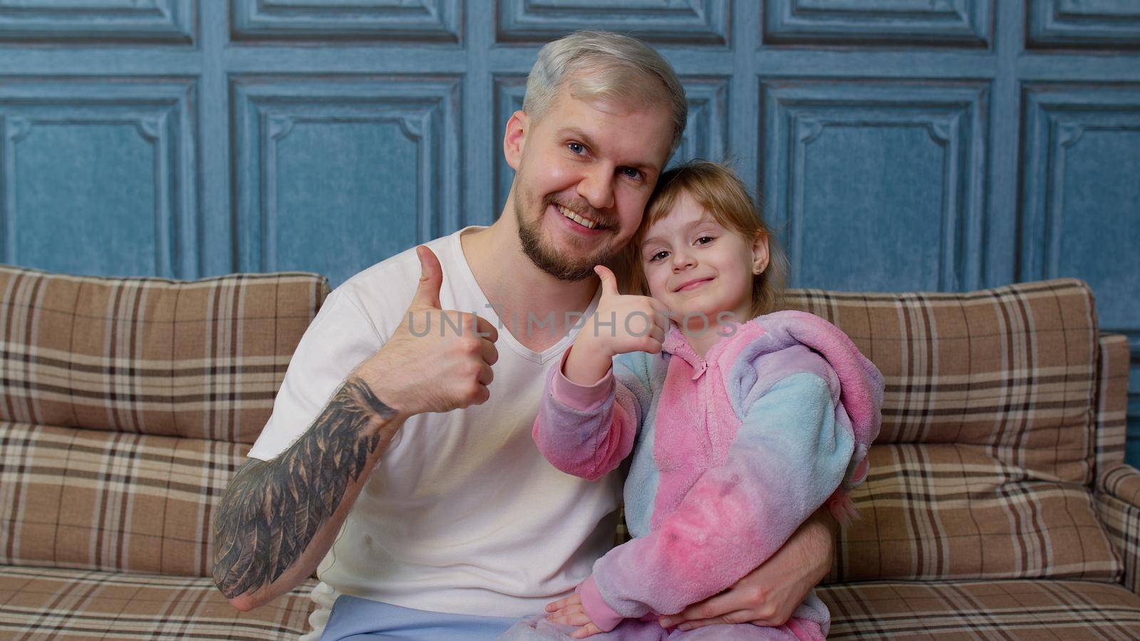 Father and little child daughter kid in pajamas sit on couch in living room smiling, looking at camera, embracing, showing thumbs up gesture together. Girl with young man. Bonding family generation