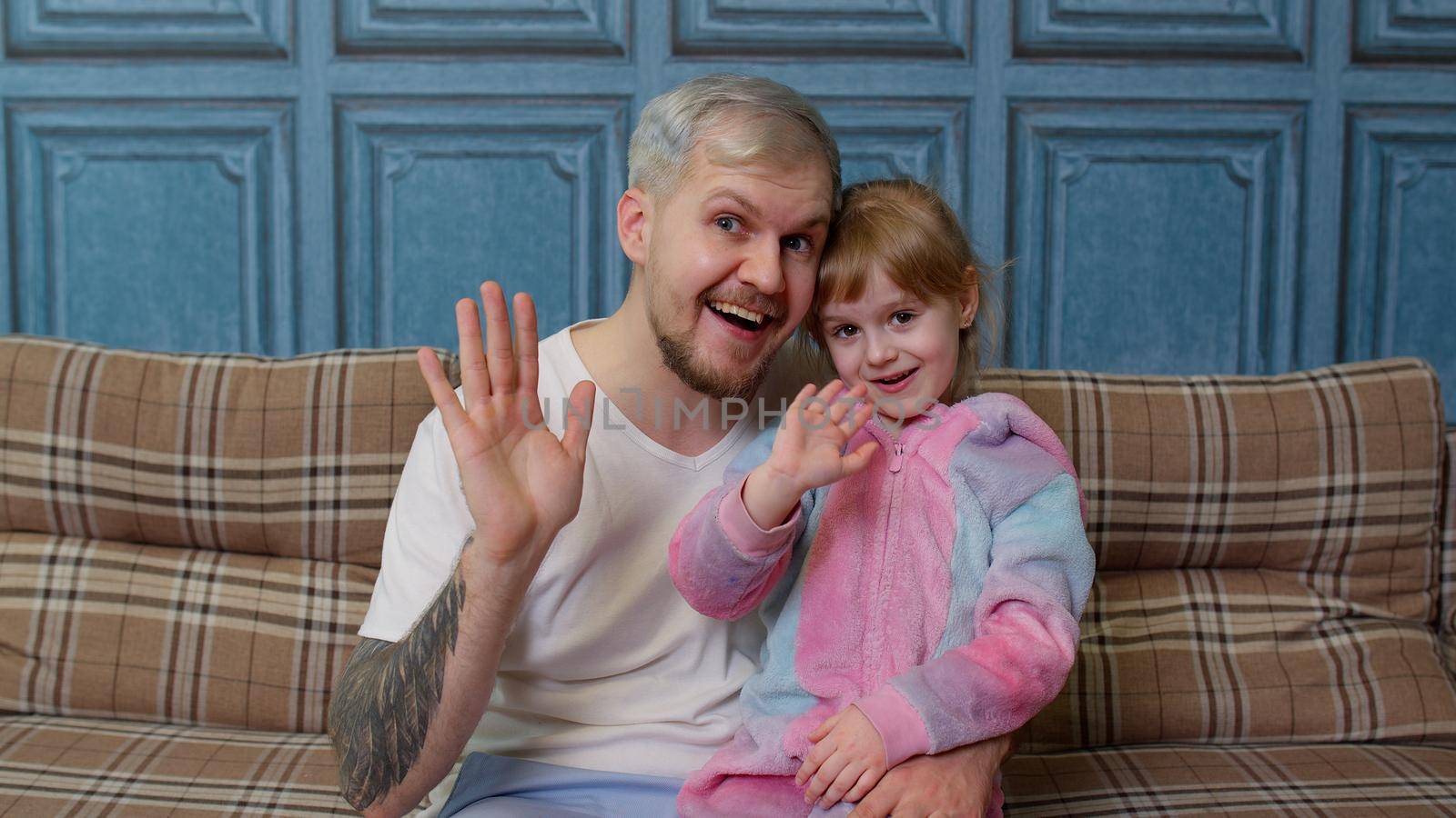 Father and little child daughter kid in pajamas sit on couch in room smiling, looking at camera, joyfully greeting, waving hi, hello, welcome together. Girl with young man. Bonding family generation