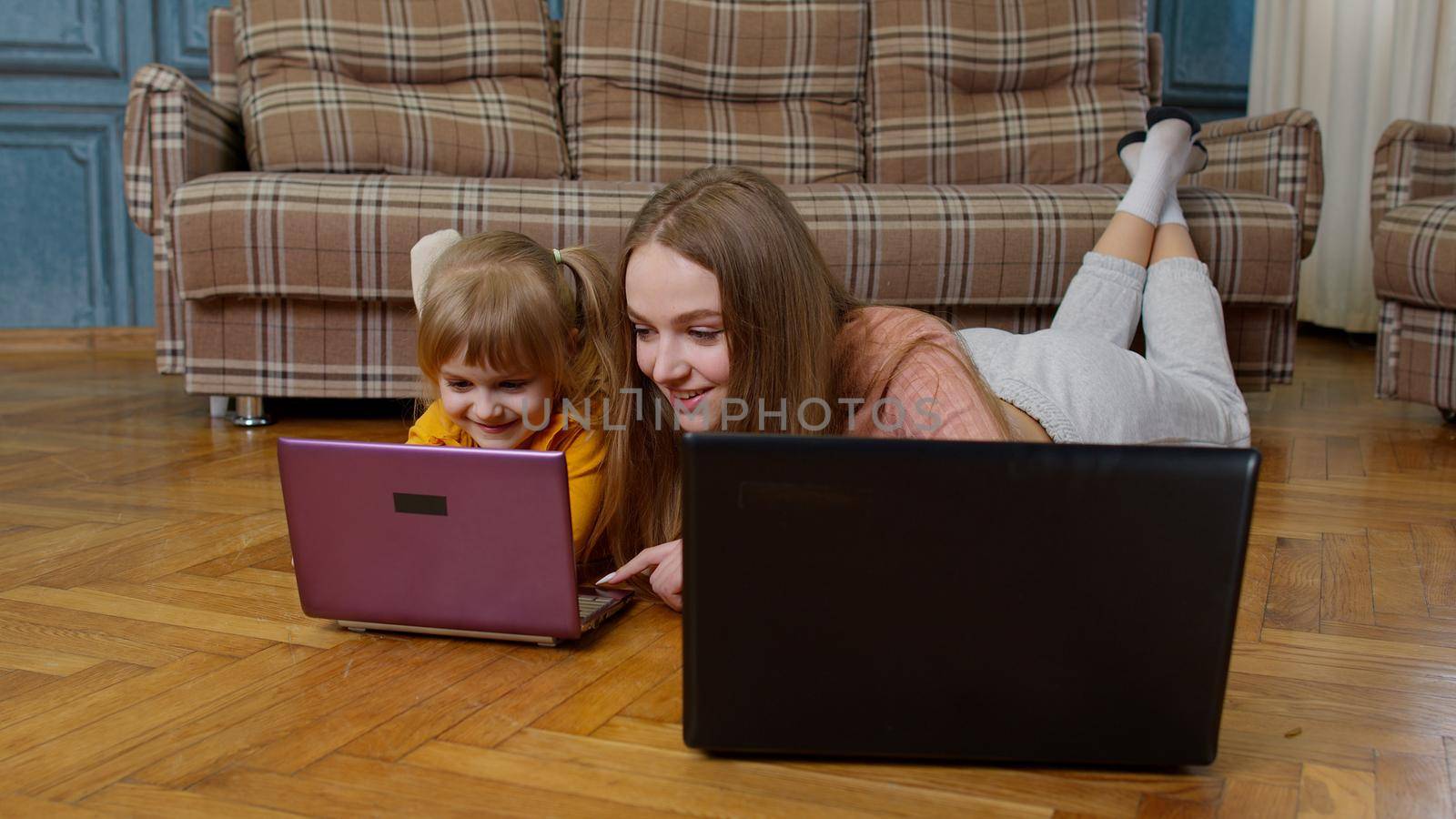 Woman nanny and child girl studying together with computer laptop, while lying on warm floor at home. Family mother and cute kid working, typing on pc, kid watching online education lessons, cartoons