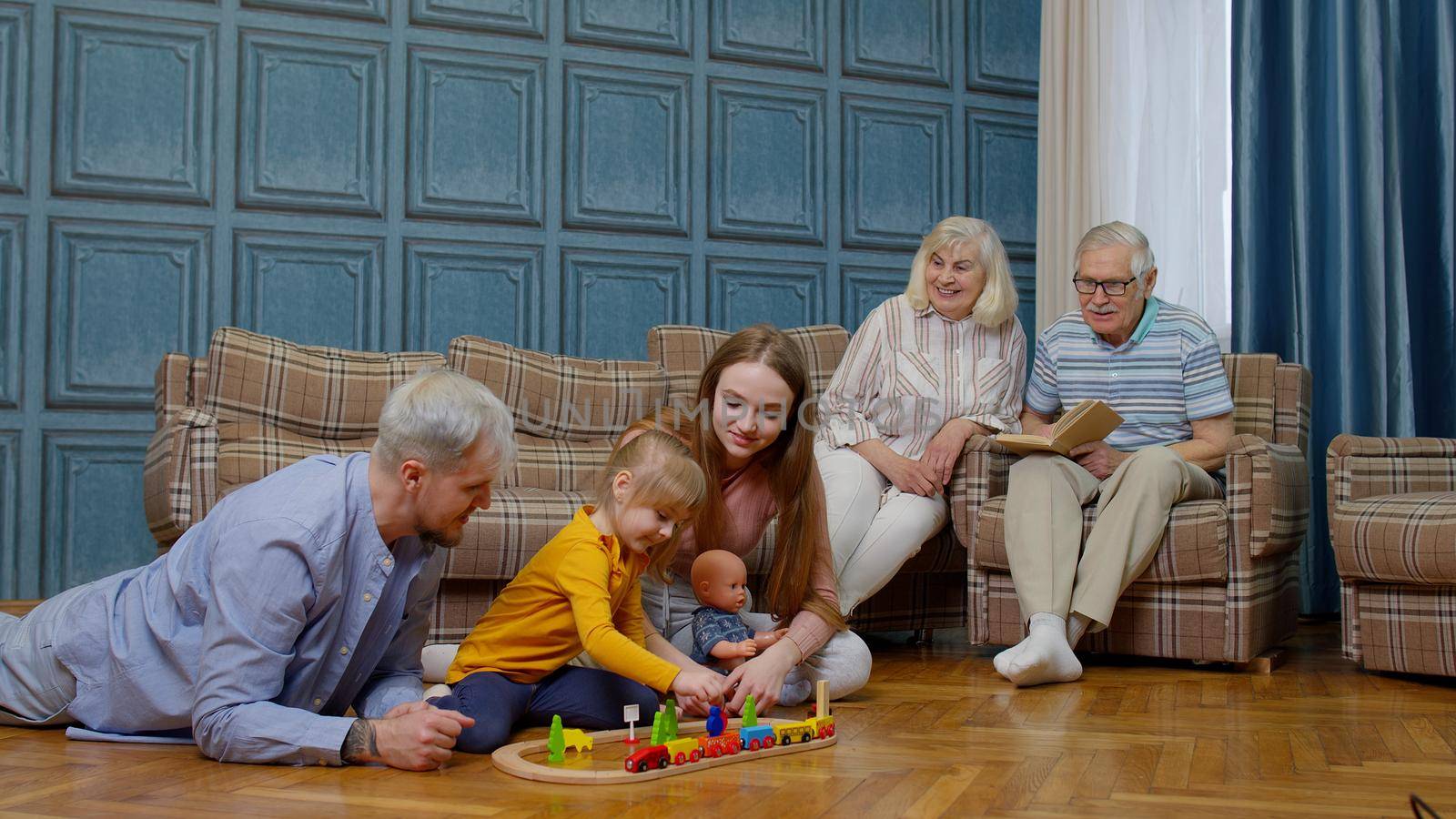 Multigenerational family leisure at home, child kid daughter playing with mother and father railway toy game on floor. Grandpagents couple relaxing talking on sofa couch in living room and having fun
