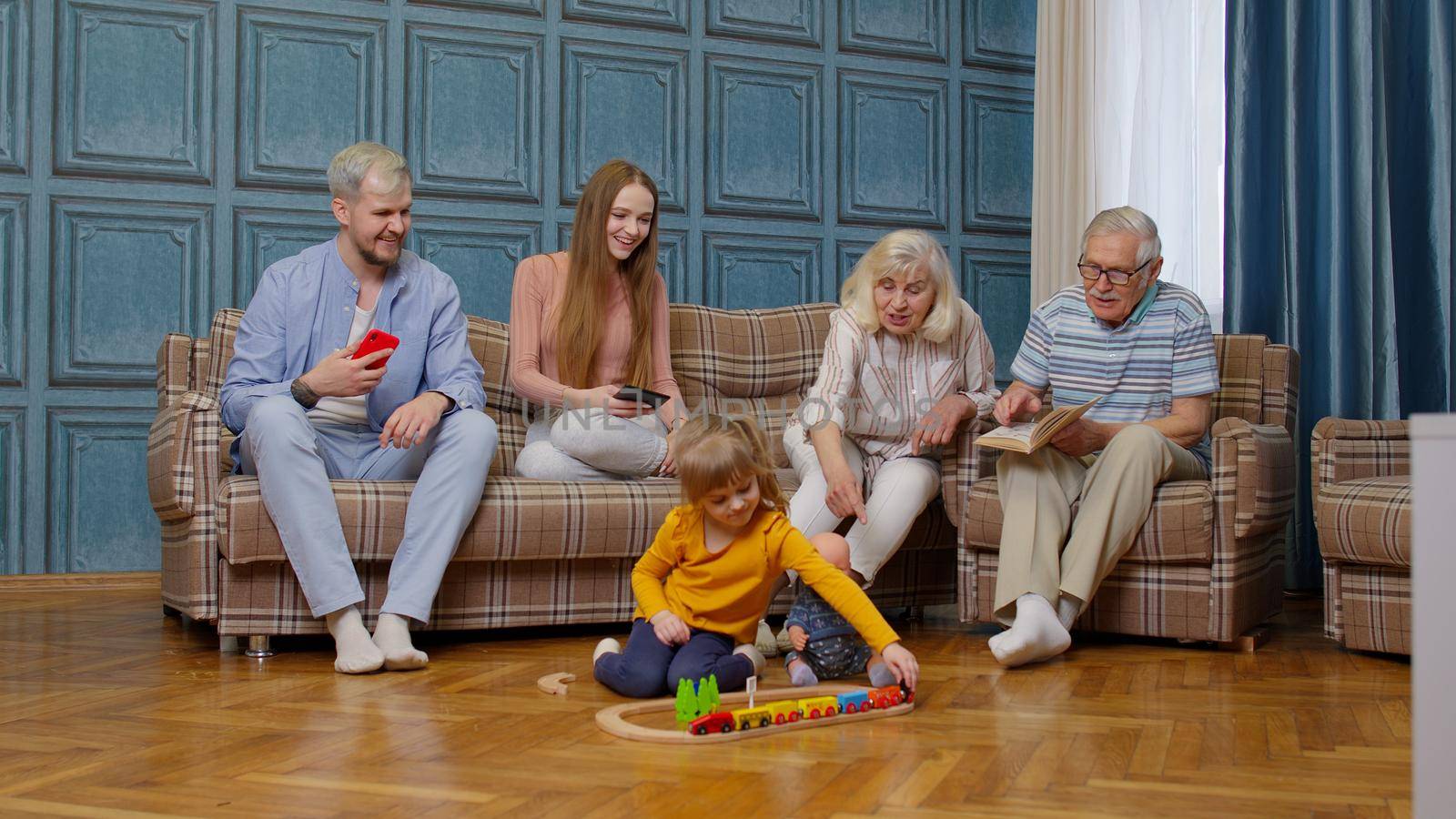Multigenerational family leisure at home, couple parents and grandpagents relaxing talking on sofa couch in comfort living room, looking at little kid child daughter playing railway toy game on floor