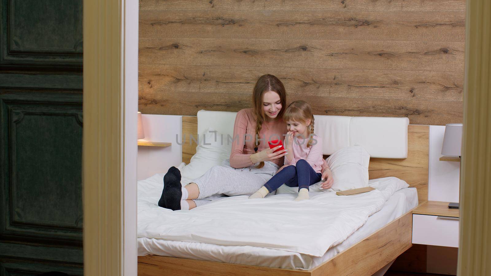 Young family of mother with joyful daughter kid girl lying on bed duvet blanket, holding cell phone, using funny mobile apps, having fun, watching cartoons, making online shopping together in bedroom