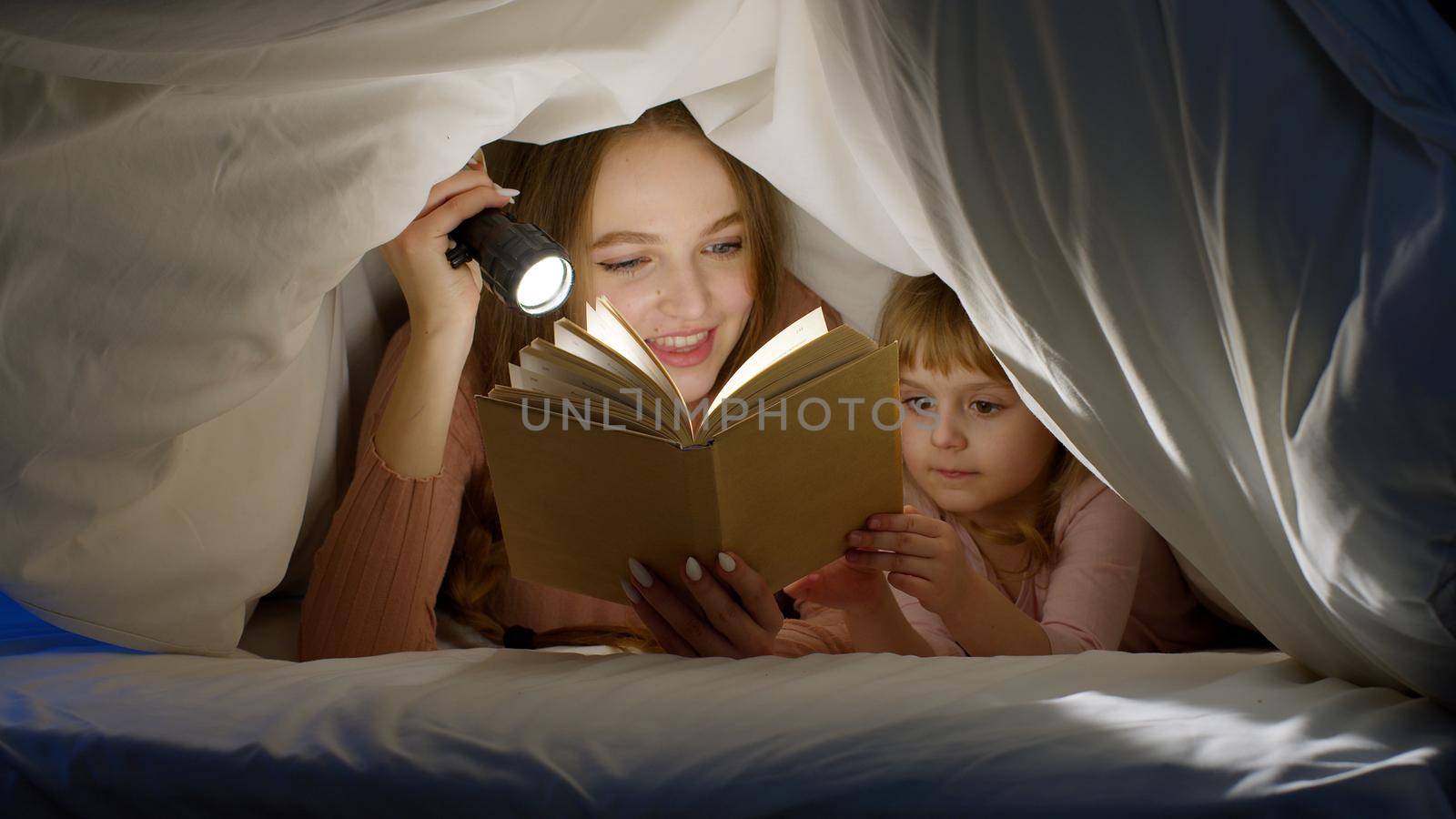 Young mother and teen daughter lying in bed at home under duvet blanket and reading bedtime stories and fairytales together with warm flashlight lamp at night. Family bonding, parenthood concept