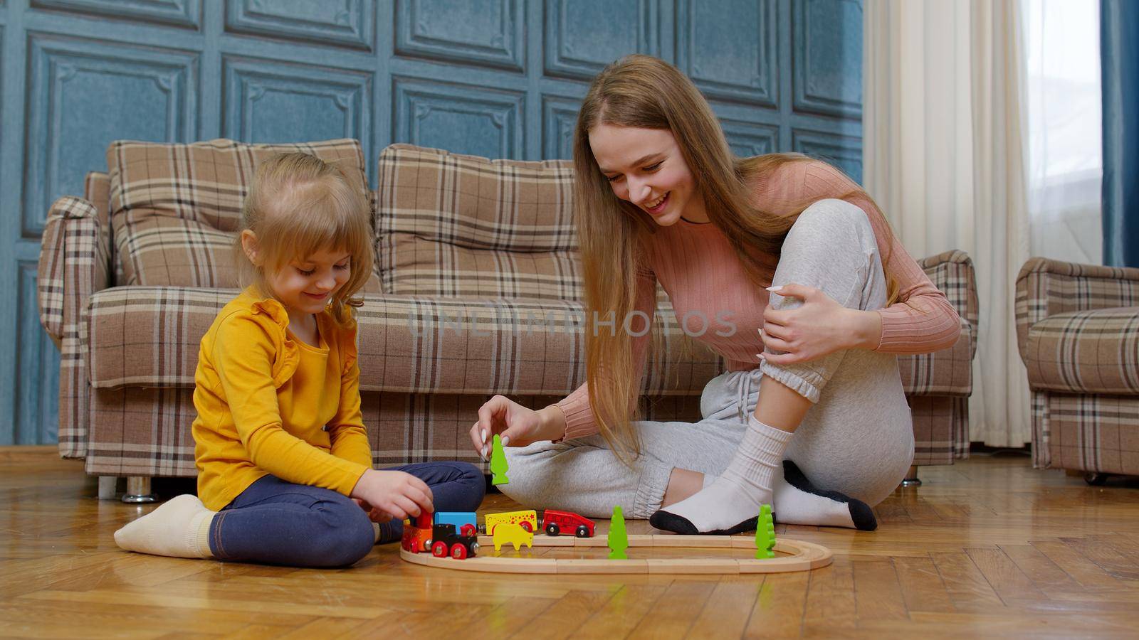 Mother with little daughter child girl riding toy train on wooden railway blocks board game. Family enjoy playtime together sit on floor in living room at home. Kid developmental game, leisure hobbies
