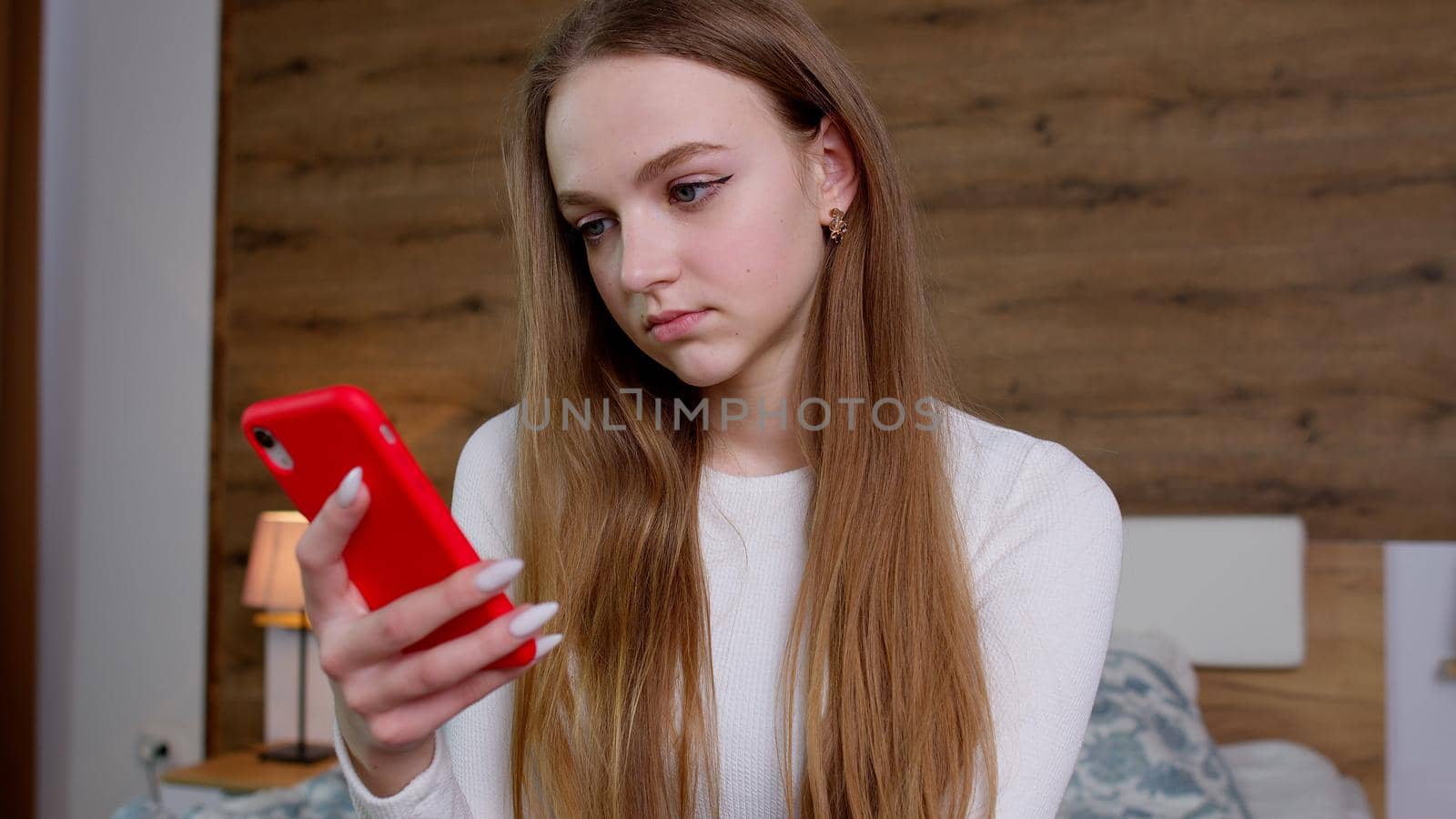 Young girl looks at screen of smartphone makes successful online order. Concentrated woman uses mobile phone and view at products on internet, browsing, surfing online while sitting on bed at home