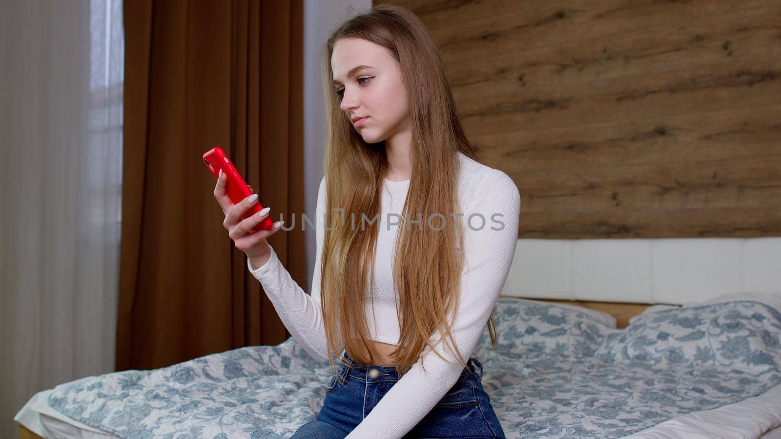 Concentrated young woman uses mobile phone and view at products on internet, browsing, surfing online while sitting on bed at home. Girl looks at screen of smartphone makes successful online order.
