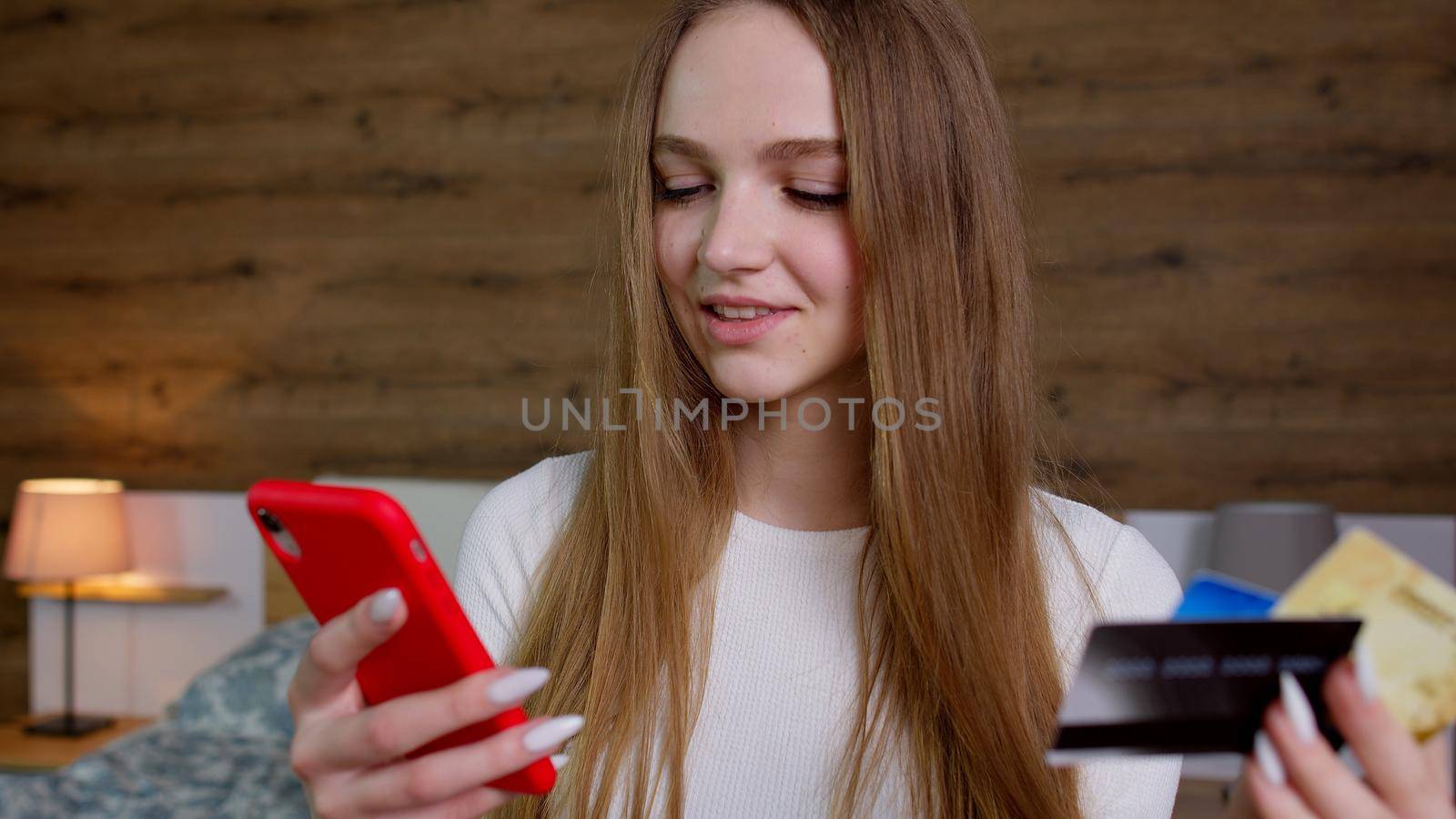 Online banking with mobile phone. Young woman sitting on bed in modern room apartment, ordering food, making shopping payments purchases transaction on Internet from bank credit card on smartphone.