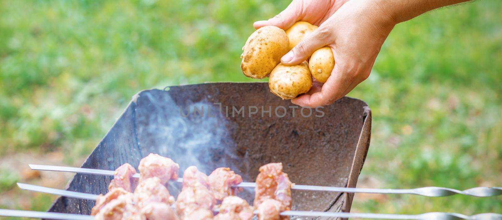 Hands of man prepares barbecue meat with potatoes on skewer by grill on fire outdoors. Concept of lifestyle rustic food preparation