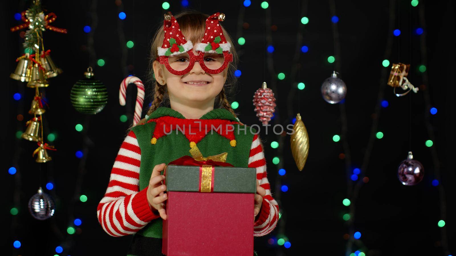 Lovely teen kid girl in Christmas elf Santa helper costume isolated on black background with garland. Child giving present gift box to camera, smiling sweetly. People New Year holidays celebration