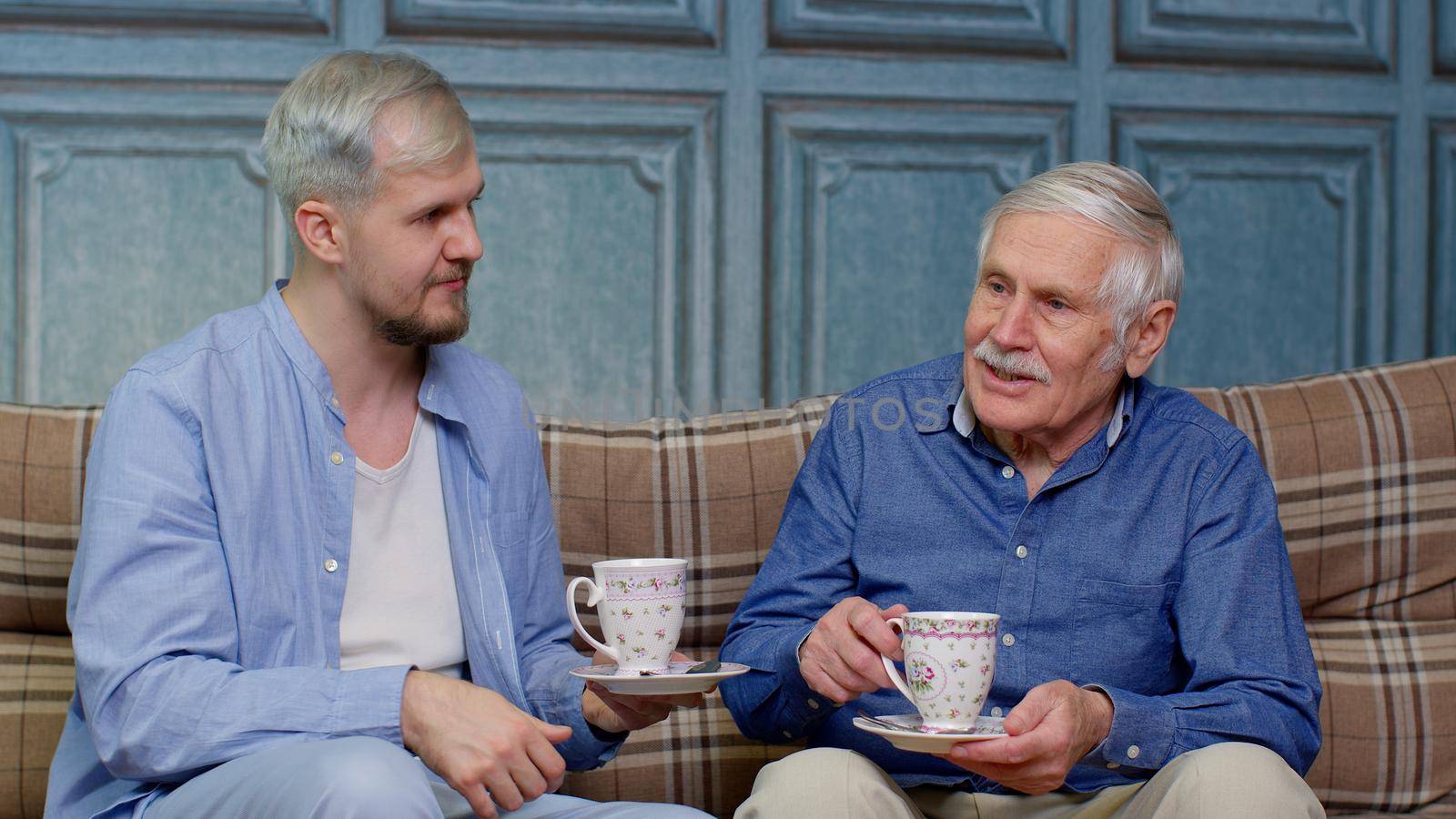 Different male generations family of senior grandfather and adult grandson relaxing on sofa, enjoying conversation, sharing problems, drinking tea. Son-in-law explaining difficulties to father-in-law