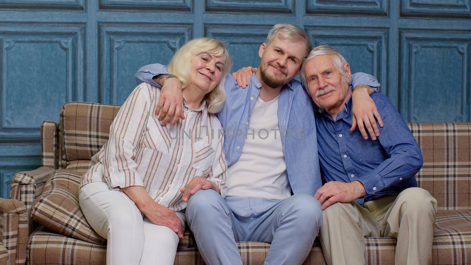 Portrait of happy generation family of senior grandparents with adult son embracing having fun looking at camera bonding at home, loving mother father. Man hugging grandmother and grandfather on sofa