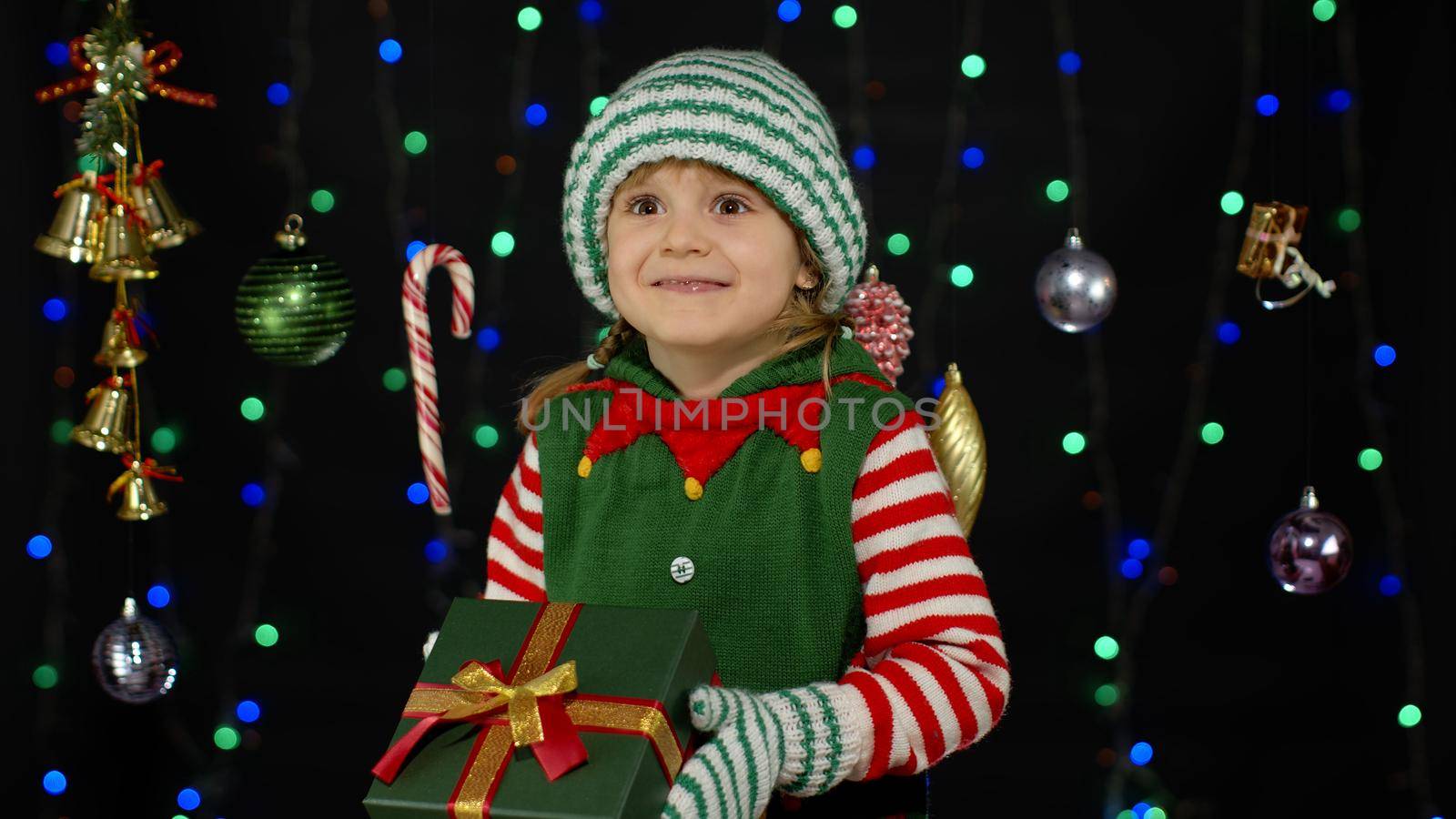 Kid child in Christmas elf Santa Claus helper costume holding present surprise red gift box with ribbon isolated on black background. People New Year holidays celebration