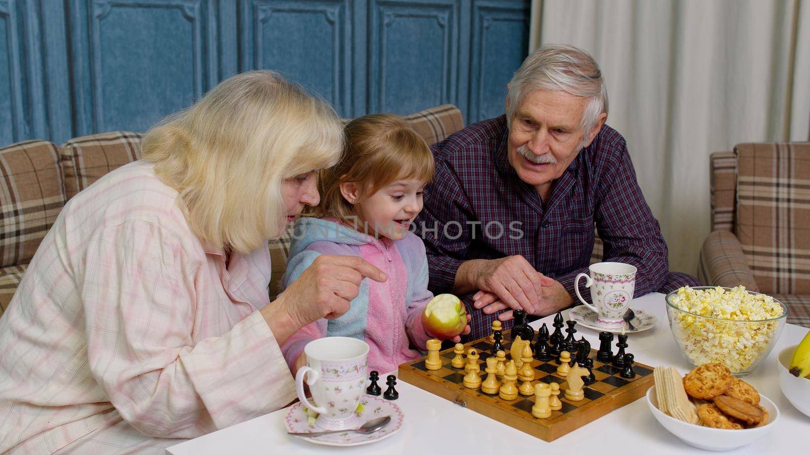 Mature grandmother grandfather with child girl grandchild playing chess game with on table in living room. Happy senior couple grandparents and granddaughter relaxing, enjoying board game at home