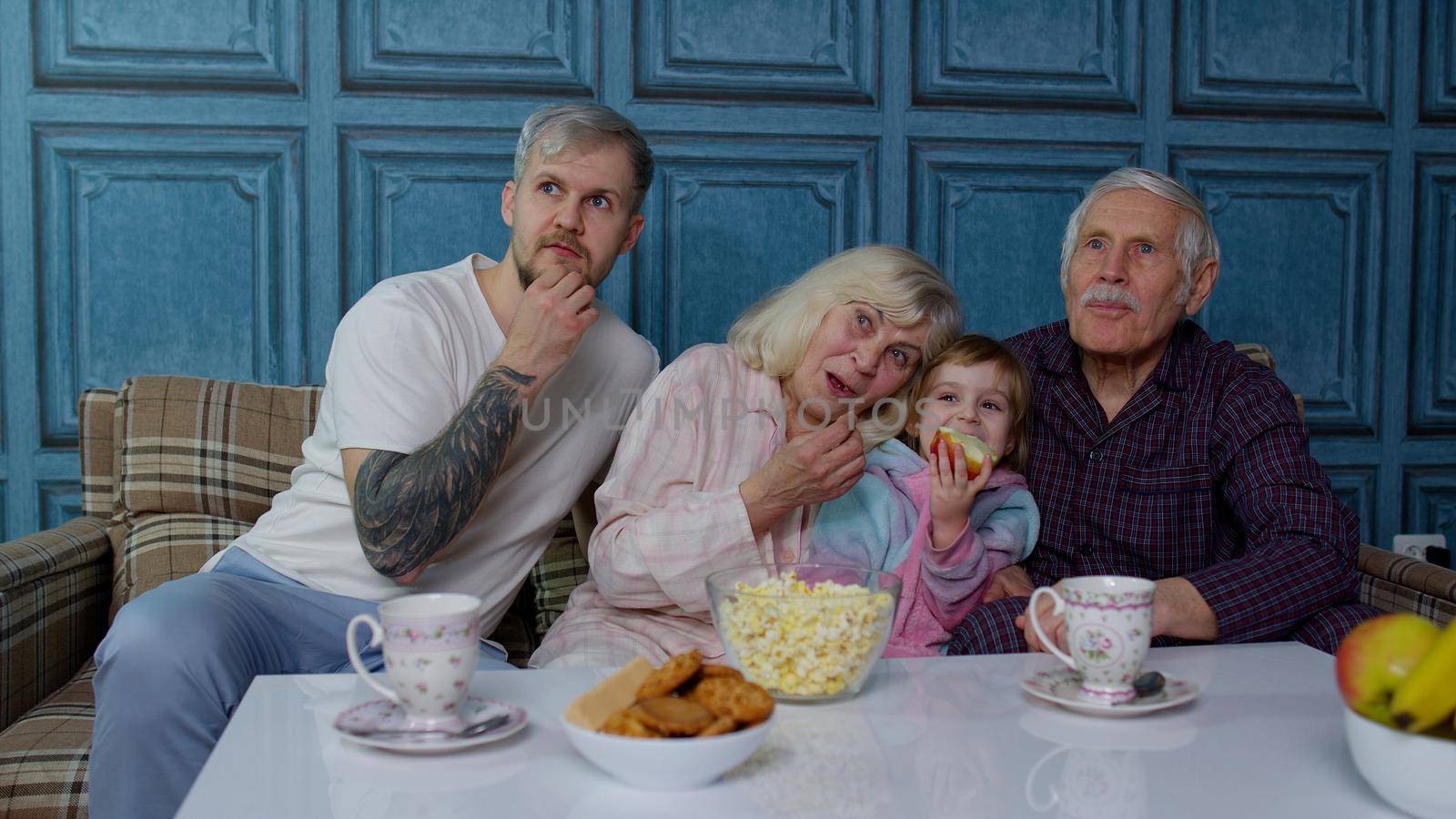 Multigenerational family watch TV with girl kid at home. Senior old grandfather and grandmother relaxing with young granddaughter sitting on sofa in living room getting scared watching horror movie