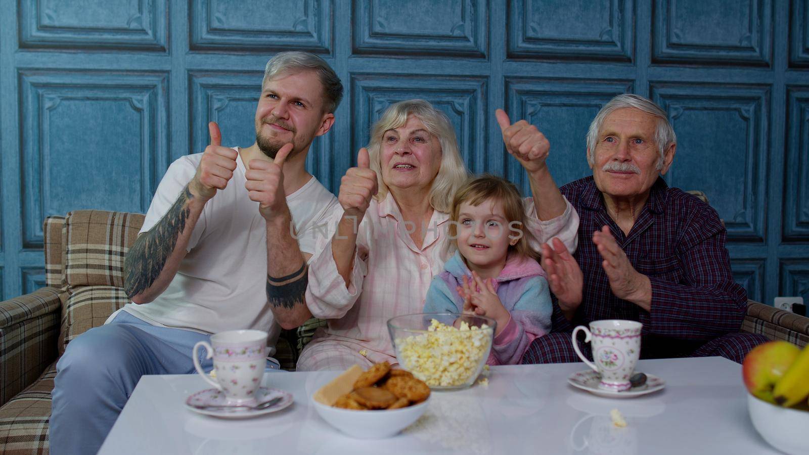 Multigenerational happy family laughing, watching comedy cartoons television movies eating popcorn at home. Senior couple grandfather and grandmother relaxing with adult son and girl kid granddaughter