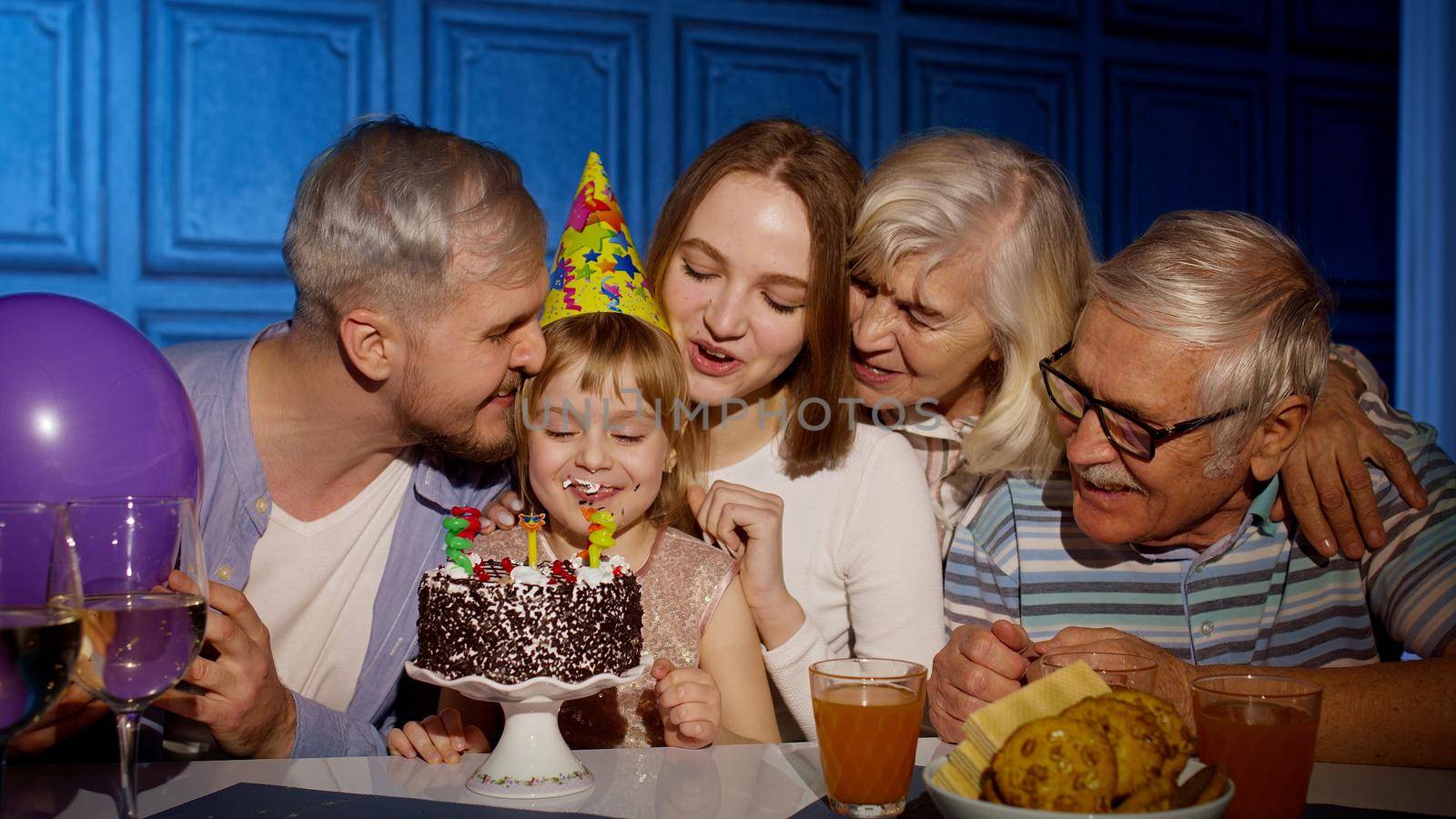 Adorable child girl kid eating cake with candles, making wish, having fun, celebrating birthday party with multigenerational family. Mother father grandparents making surprise, clapping hands together