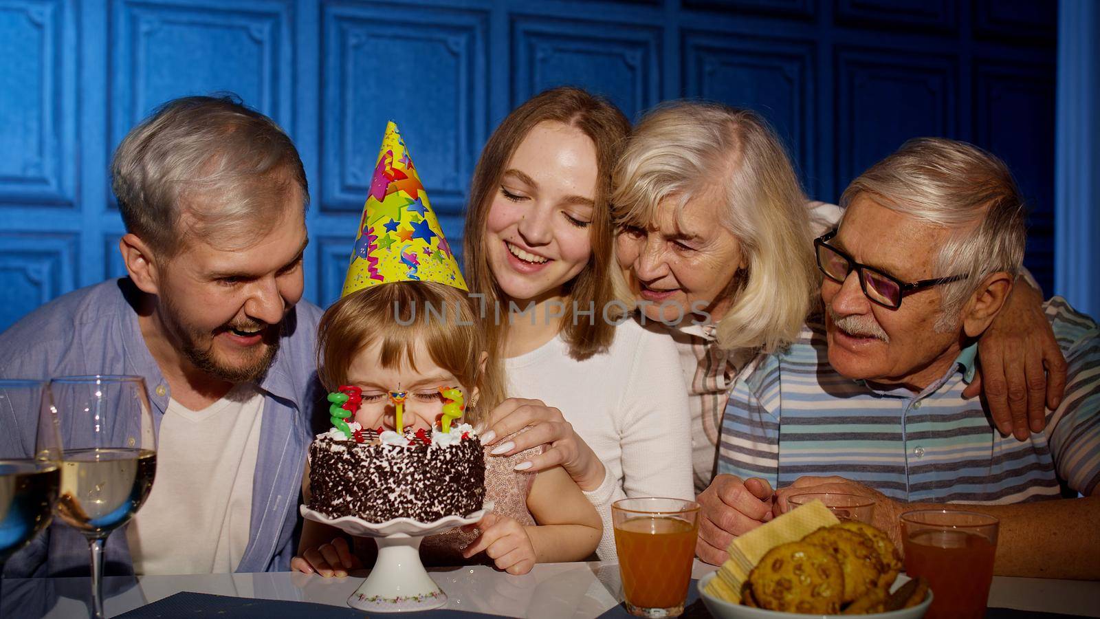 Adorable child girl kid eating cake with candles, making wish, having fun, celebrating birthday party with multigenerational family. Mother father grandparents making surprise, clapping hands together
