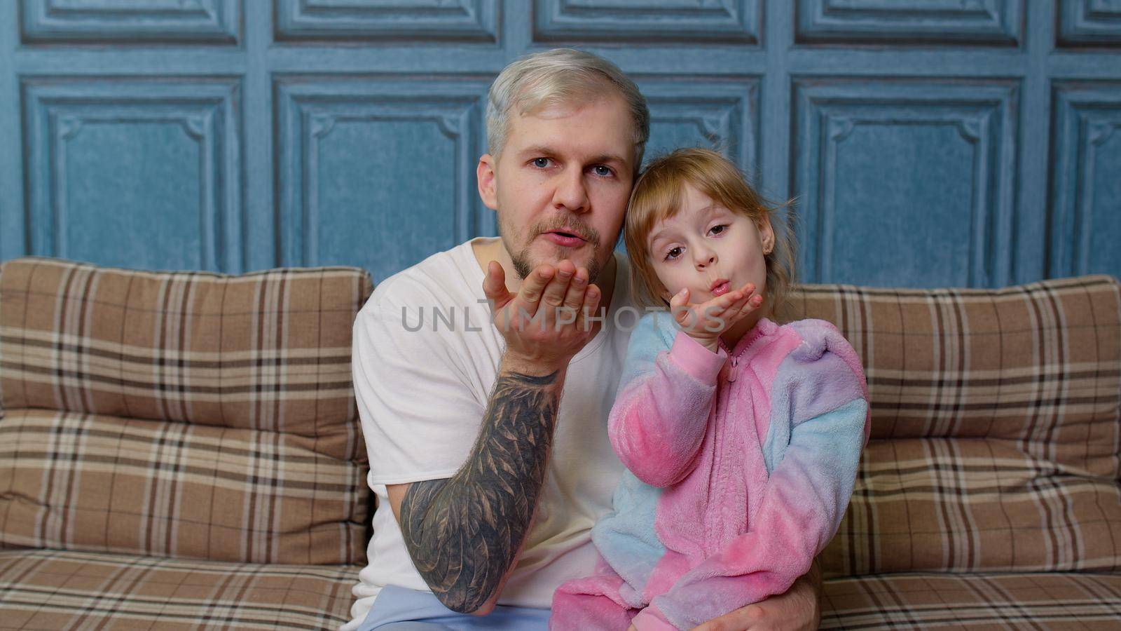 Father and little child daughter kid in pajamas sit on couch in room smiling, looking at camera, joyfully blowing sending an air kiss together. Girl with young adult man. Bonding family generation