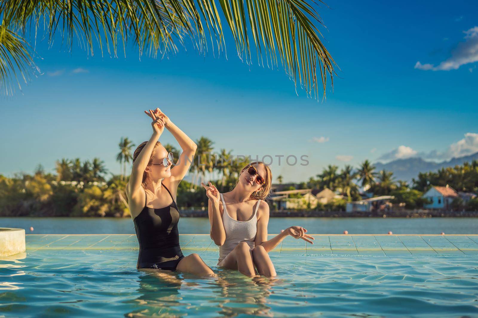 Two beautiful young woman sitting by the poolside of a resort swimming pool during summer holiday. Party, tourism in tropical countries.